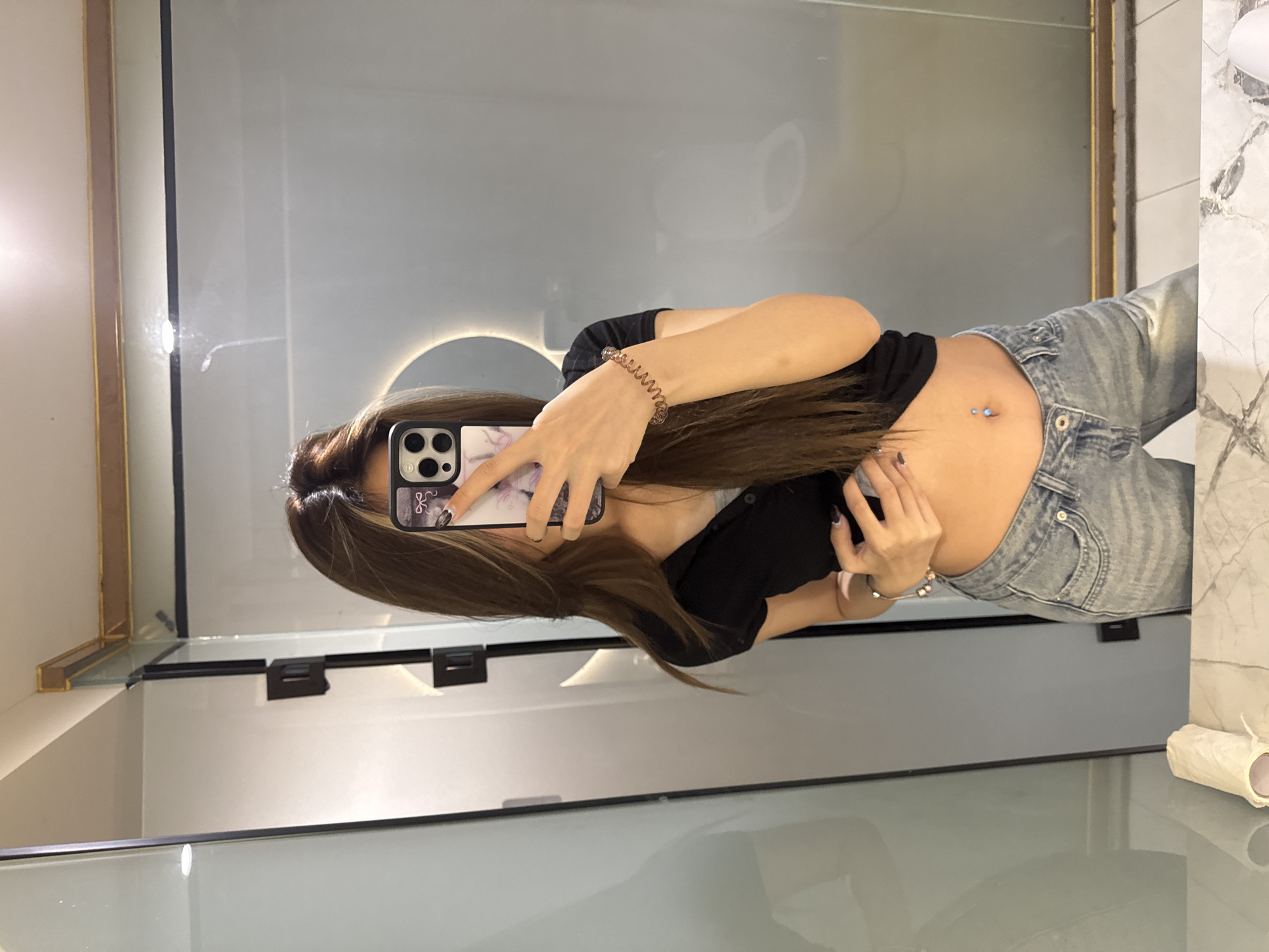 A young woman is taking a selfie in a bathroom. She is wearing a black shirt and light blue jeans, which sit low on her waist, exposing her belly button, where there is a piercing. She has long brown hair. She is holding a smartphone with a decorative case. The bathroom features a large mirror, a modern sink, and marble-style wall tiles. The location is likely a private residence in Yuzhong District, China.