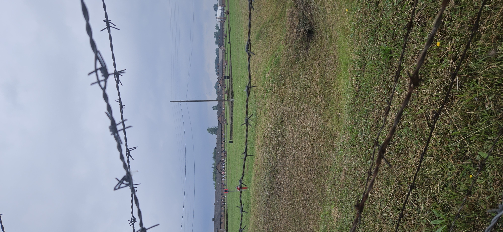 The image shows a view of the Auschwitz I concentration camp in Brzezinka, Poland. In the foreground, barbed wire fences are visible, creating a sense of separation and confinement. Beyond the fences, there are rows of barracks, characteristic of concentration camp architecture, with several figures, likely visitors, walking in the open space between them. The sky is overcast, contributing to a somber atmosphere. A red and white sign, possibly indicating a historical marker or informational display, is present in the middle ground. The overall scene depicts a historical site of significant importance, conveying a stark and solemn impression.