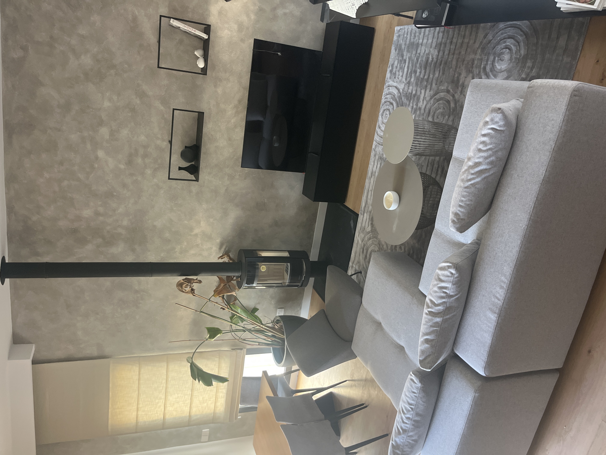 This is a medium shot, eye-level, interior photograph taken in a modern living room. The scene is well-lit, with natural light coming from a window on the left side of the image. The room features a light grey sectional sofa in the foreground, with several throw pillows. A grey rug with a circular pattern is placed on the wooden floor in front of the sofa. Two circular coffee tables are situated on the rug, one with a white candle. A large, modern black television is mounted on the wall above a black media console. To the left of the television, a black wood-burning stove stands on a black hearth. A large potted plant with green leaves is positioned next to the stove. In the background, a dining table with chairs is visible, and a window with a light-colored Roman blind is present. The walls are a textured, neutral grey color, and two black floating shelves are mounted on the wall above the television. The scene appears to be located in Agios Athanasios Municipality, Cyprus, based on the context provided. There are no people visible in the image.