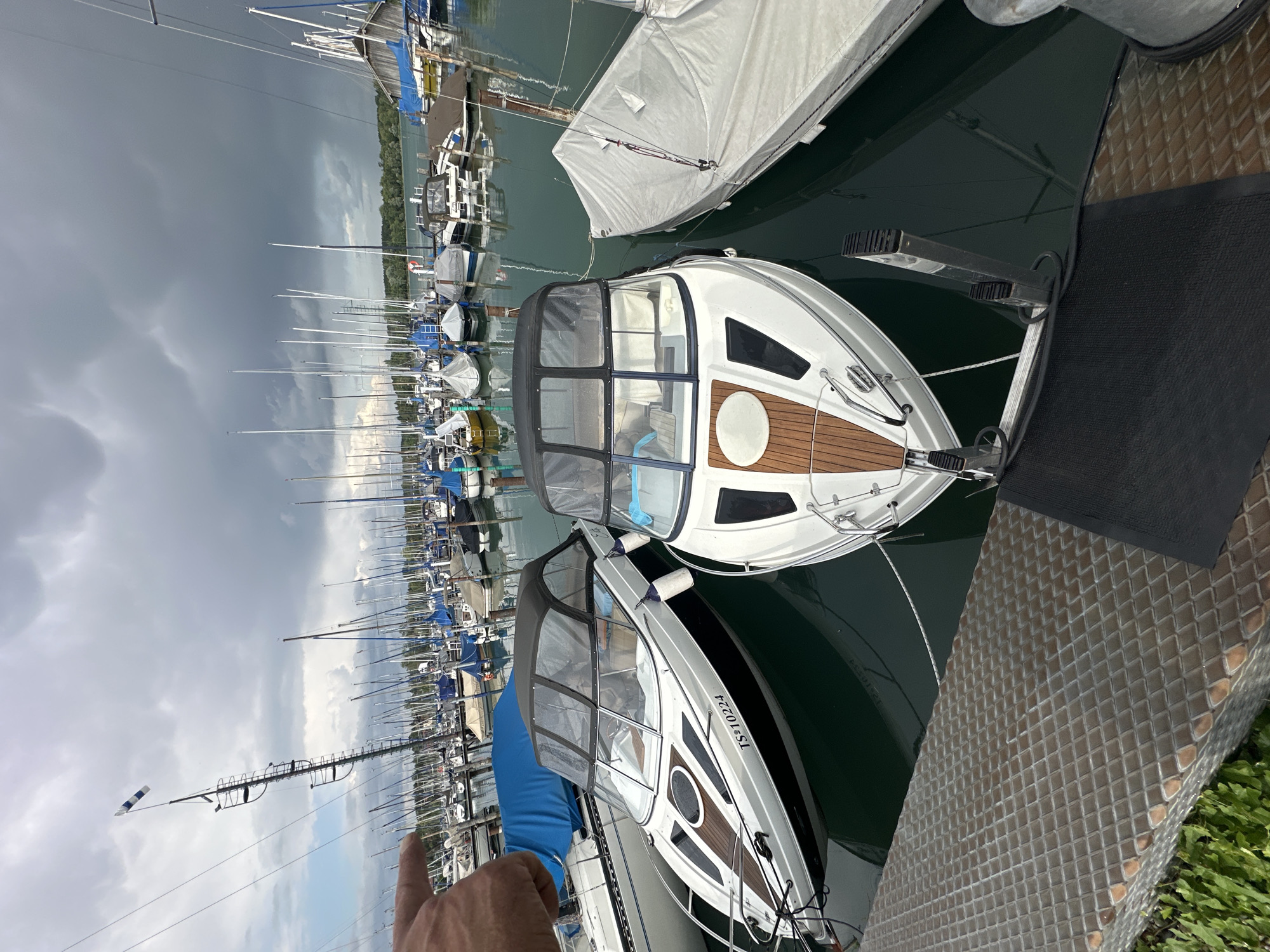 A wide-angle, eye-level shot captures a marina on a cloudy day, likely in Chiemsee, Germany, given the presence of numerous sailboats and the distinctive German license plate "TS" visible on a motorboat. In the foreground, a textured, metallic pier juts out into the dark green water. Two white motorboats with black trim and canvas tops are docked side-by-side. The boat on the left displays the license plate "TS 10224." Adjacent to these motorboats, a sailboat is covered with a large, light-colored tarp. Behind these boats, a dense collection of sailboats with tall masts, many with blue covers, fills the frame, stretching towards a tree-lined horizon under a dramatic, overcast sky. A hand with a pointing index finger is visible in the upper left corner, its placement suggesting it is interacting with the scene, possibly indicating something in the distance.