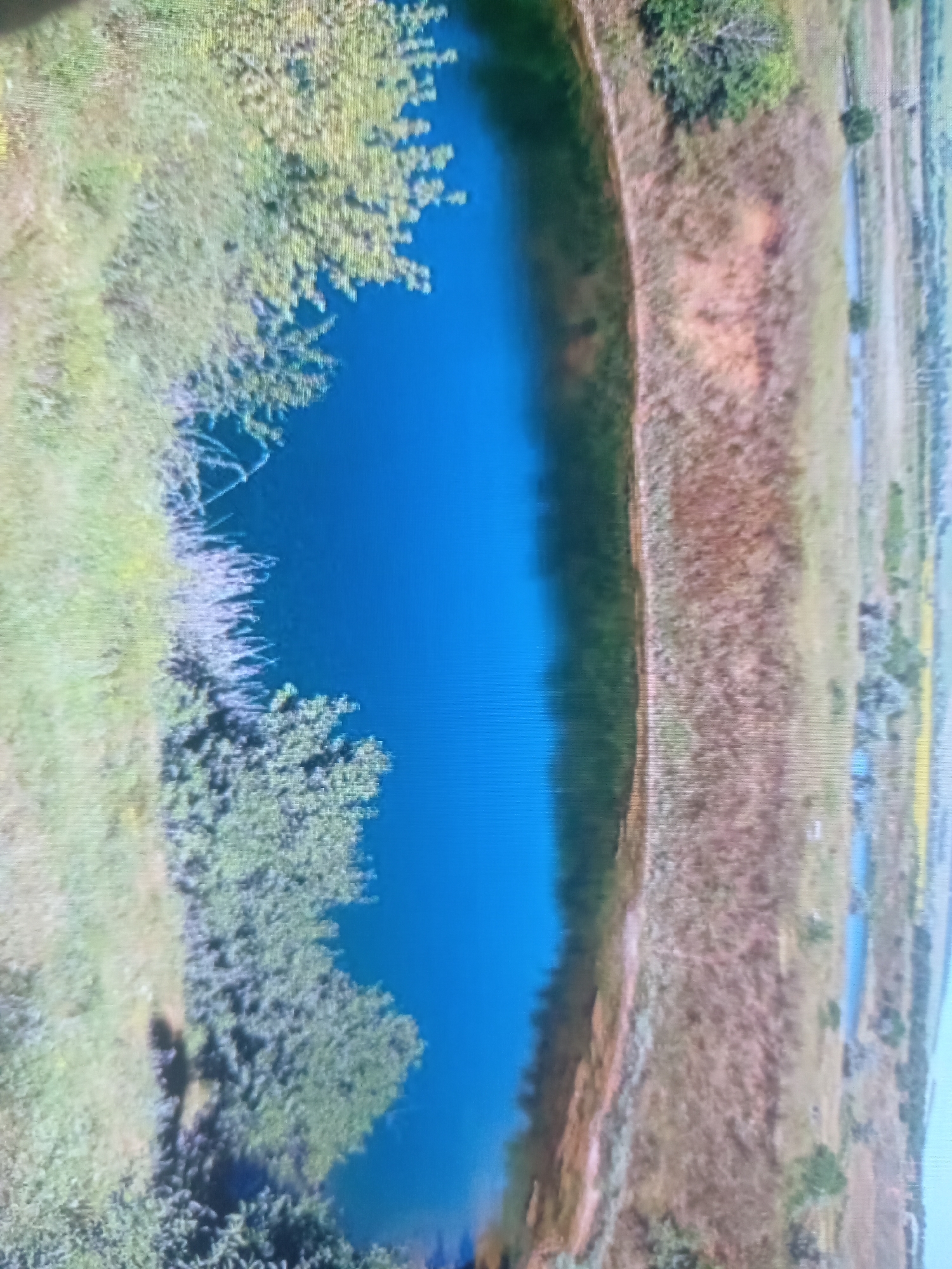 The image is an aerial view of a body of water with a distinct blue hue, likely a pond or lake. The water's edge is bordered by vegetation on the left, consisting of trees and lighter-colored brush. The right side of the image shows a dry, barren landscape with patches of reddish-brown soil and sparser vegetation, appearing to be a field or savanna. Several narrow, lighter-colored channels or ditches are visible running parallel to each other in the distance, suggesting an agricultural or water management area. The image is rotated 90 degrees clockwise. There are no people or specific objects of interest clearly discernible in the scene. The overall environment appears to be rural or undeveloped.