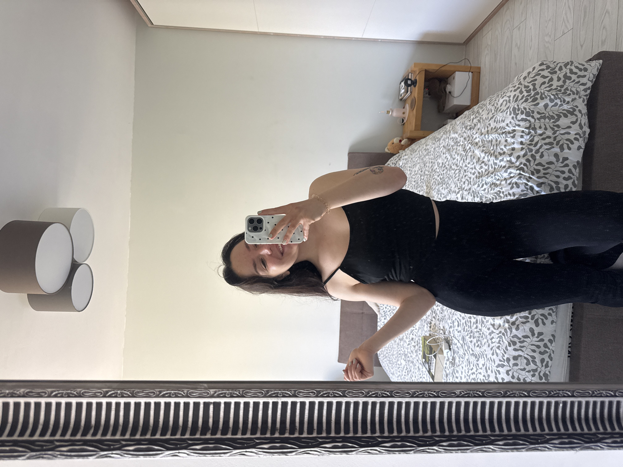 An indoor mirror selfie captures a young woman with long dark hair and a light complexion, seen smiling faintly. She is wearing a black spaghetti-strap tank top and black fitted pants, with a small, indistinct tattoo visible on her upper right arm. She holds a white smartphone with a black polka-dot case, featuring prominent camera lenses, in her right hand. Her fingernails are painted a light teal color.
The mirror's decorative frame, visible at the bottom, is dark-toned with textured striped and wavy patterns. The room reflected in the mirror is a brightly lit bedroom, suggesting daytime. The walls are light-colored, appearing as a pale cream on the right and a lighter shade on the left. On the left, a wall-mounted light fixture with three stacked cylindrical shades (two grey, one white) is visible. On the right, a bed features a grey and white duvet cover with a dense, leaf-like pattern. Next to the bed, a wooden bedside table holds a small pink fan, a brown teddy bear, and some cables. The floor appears to be light-colored wooden planks. No discernible text is visible in the image. The location context provided is Burgas, Bulgaria.