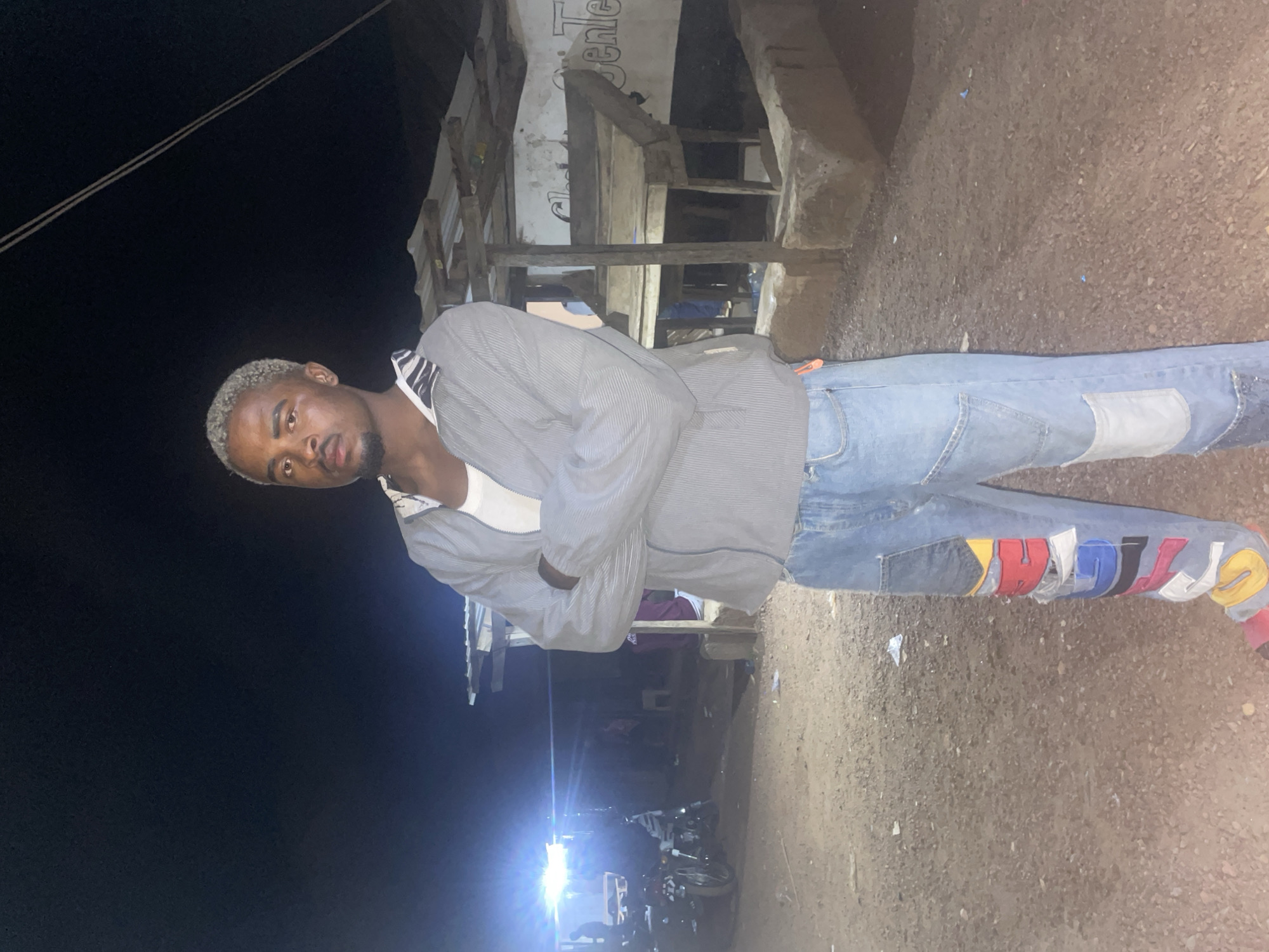 A man stands in front of a building at night in Durimi, Nigeria. The man has blond hair and wears a gray striped shirt and blue jeans with colorful patches. He stands with his arms crossed. A motorcycle is visible in the background, illuminated by headlights. The ground is dirt.