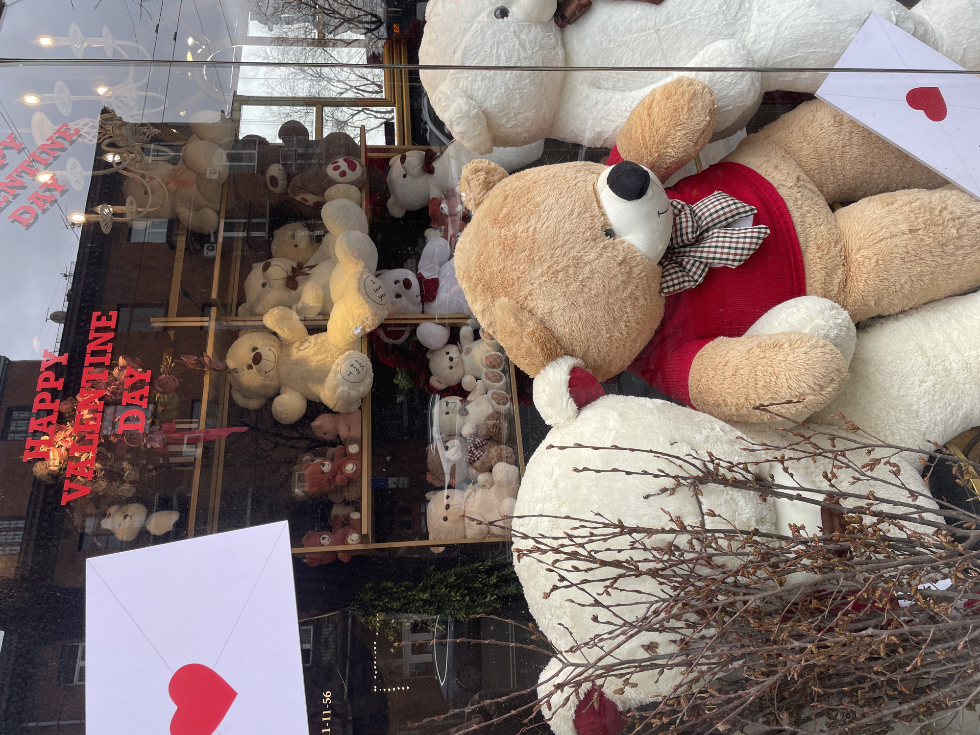 This image displays a retail storefront window in Yerevan, Armenia, featuring a Valentine's Day promotional arrangement. Numerous plush teddy bears of assorted sizes and colors are positioned throughout the display, both on wooden shelving units and directly on the floor of the window. In the foreground, a large, light brown teddy bear wearing a red long-sleeved shirt and a black-and-red plaid bow tie is seated prominently. To its right, a large white teddy bear with a brown bow tie is partially visible. To its left, another large cream-colored teddy bear is partially obscured by a cluster of bare, budding branches positioned in the immediate foreground. Behind these, additional teddy bears in various shades of white, brown, and pink are arranged on tiered wooden shelves. Several teddy bears feature "I love you" text on their paws. Red cutout letters spelling "HAPPY VALENTINE DAY" are affixed to the interior of the glass window. A large white paper envelope with a red heart is attached to the upper-left glass, angled downwards, and a smaller similar envelope is visible at the bottom-right. The window glass reflects an exterior urban environment, including an overcast sky, bare deciduous trees, a dark-colored parked vehicle, and brick buildings with windows. The reflection of an ornate multi-bulb chandelier is also visible in the upper portion of the glass.