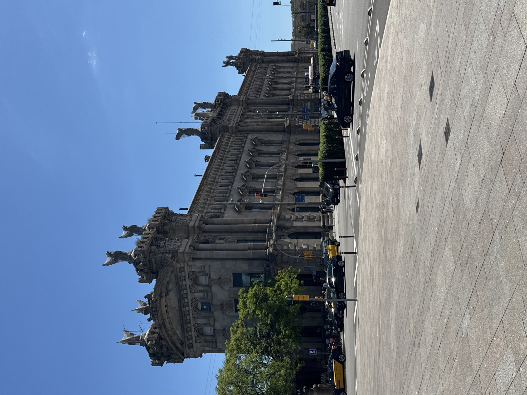The image shows a street scene in Barcelona, Spain. A grand, ornate building with multiple statues on its roofline dominates the left side of the frame. A line of parked and moving cars is visible on the right, adjacent to a wide, paved sidewalk. The sky is clear and blue. Taxis, traffic lights, and pedestrians can also be seen in the scene.