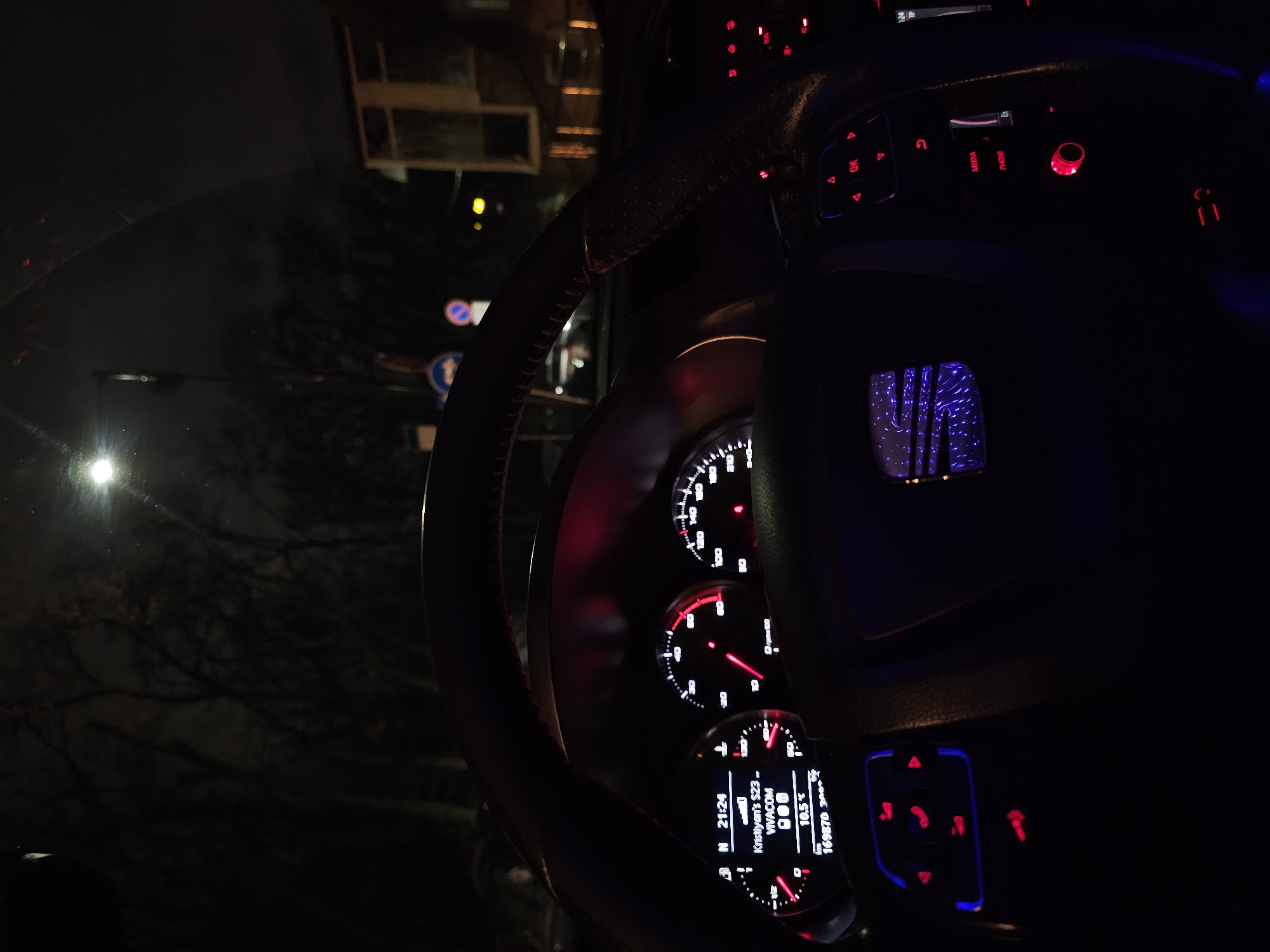 Here is a concise description of the image:
The image shows the interior of a car, likely in Sofia, Bulgaria, based on the Cyrillic alphabet on the dashboard display. The focus is on the steering wheel, dashboard, and instrument cluster, all illuminated in red and blue lights. The instrument cluster displays information such as time, temperature, and radio station name. Outside the car, a street scene with a street lamp and buildings is visible through the windshield.