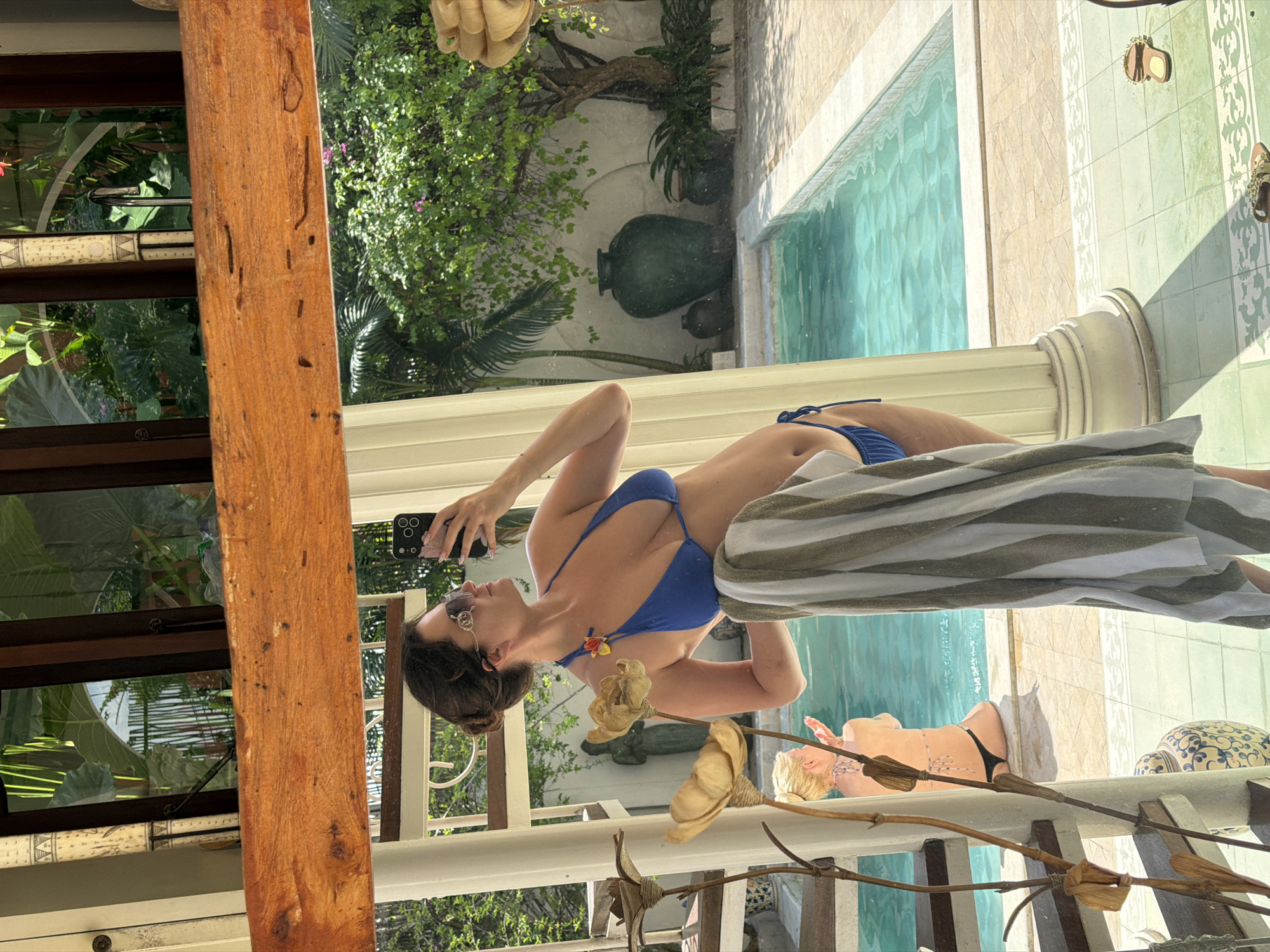 The image displays a complex scene, primarily captured as a reflection, and is oriented vertically, rotated 90 degrees clockwise. In the center, a woman in a blue two-piece bikini is holding a black smartphone with multiple cameras, appearing to take a selfie. She has dark hair tied back, wears sunglasses, and a necklace with a small floral detail.
The foreground of the reflection features a striped towel (white and light grey/green) draped over a white balustrade. This balustrade is adorned with dried, light-colored floral elements and wooden sticks. A prominent, weathered light brown wooden pillar stands on the left side of the reflection.
The reflected background reveals a sunlit outdoor area with a light blue swimming pool, surrounded by white and light green patterned tiles. Another woman, with blonde hair and possibly tattoos, is seen lounging at the pool's edge, wearing a black bikini bottom. Nearby, a pair of light-colored sandals and a patterned ceramic stool are visible.
Beyond the pool, a vibrant tropical landscape unfolds with lush green foliage, plants, and trees, along with several large, dark green decorative pottery pieces. White architectural elements, including columns and textured walls, complete the scene. The bright sunlight indicates it is daytime with clear weather. The overall setting suggests a luxurious and relaxing tropical resort or villa, consistent with a location like North Kuta, Indonesia. No discernible text is visible in the image.