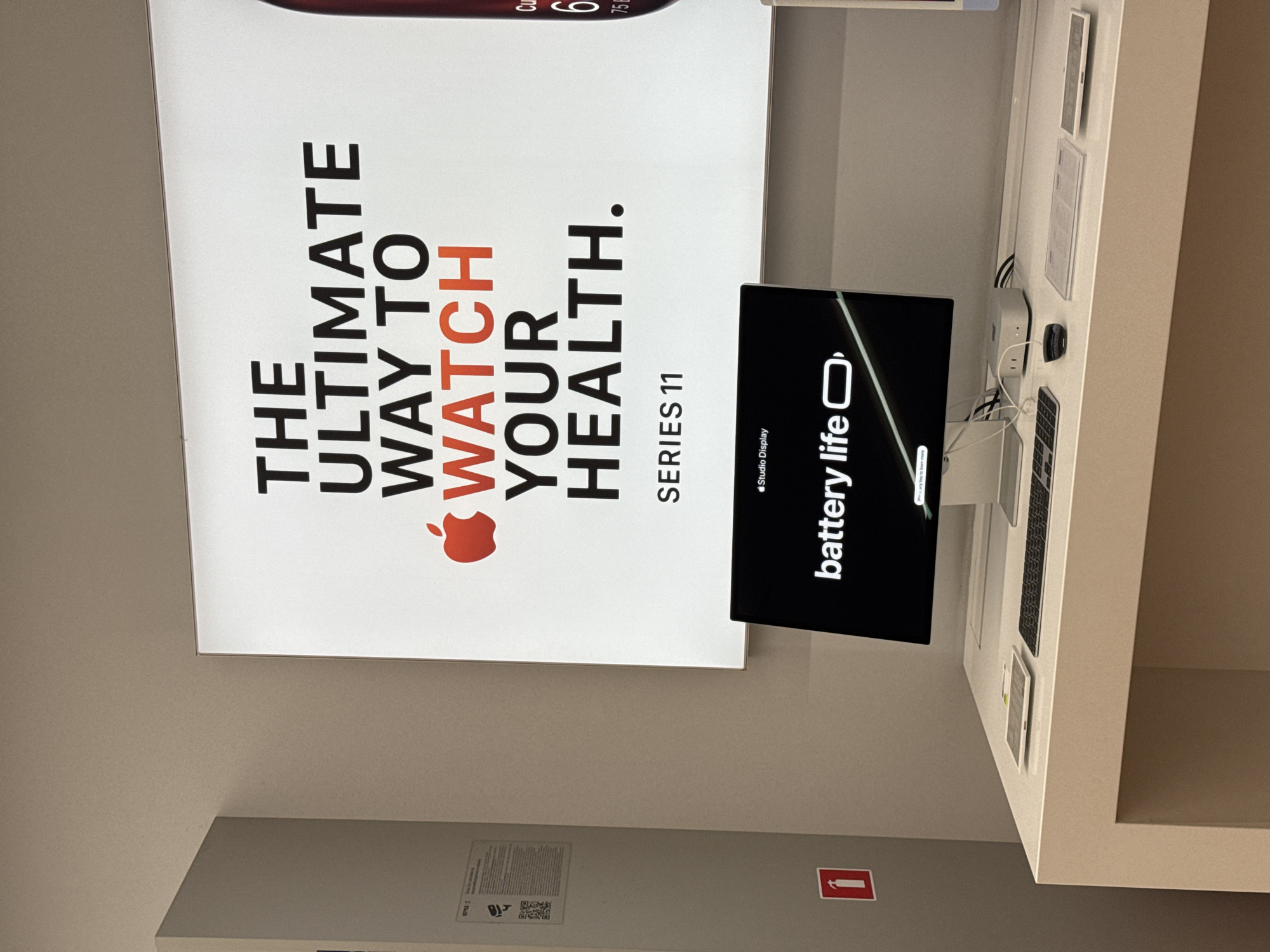 This indoor photograph, taken in Sofia, Bulgaria, depicts a section of an electronics store, likely featuring Apple products. The scene is dominated by a large, vertically oriented, backlit sign on the left wall advertising the Apple Watch. The sign's main text reads: "THE ULTIMATE WAY TO WATCH YOUR HEALTH." below which is the red Apple logo and "SERIES 11." Part of what appears to be an Apple Watch display is visible at the very top of the sign, showing "Cu 6 75," possibly indicating metrics like calories, activity rings, and heart rate or a percentage.
In front of the sign, on a white display table, is a black computer monitor, which is also oriented vertically. The monitor screen shows a dark interface with white text prominently displaying "battery life" next to a battery icon. Below this, in smaller text, "Studio Display" is visible, and even smaller text at the very bottom says "Press any key to learn more." To the right of the monitor, on the same table, a white Mac mini-like device, a black mouse, and a silver keyboard are set up, along with several informational cards or brochures. Cables are visible connecting the devices.
On the lower left wall, a small, white piece of paper with "STYLE," some text, and a QR code is affixed. Directly below this, a red fire extinguisher with a white '1' symbol is mounted on the wall. The overall lighting suggests an indoor retail environment. No people are visible in the image.