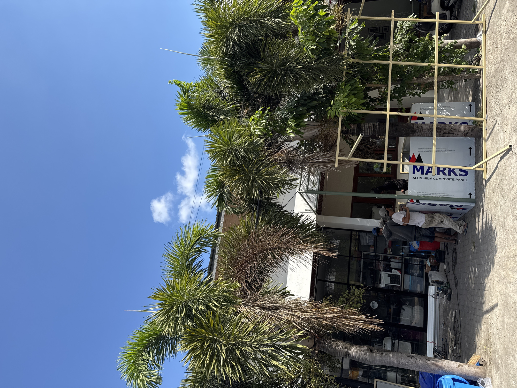 In North Kuta, Indonesia, two men are working on what appears to be a construction or renovation project outside a building. One man, wearing a grey hoodie and dark shorts, stands near a large, white panel with blue text that reads "MARKS ALUMINIUM COMPOSITE PANEL." The other man, in a light-colored shirt and tan pants, is positioned next to the same panel, facing it. They are on a paved sidewalk in front of a building with large glass windows. Several palm trees are visible in the foreground, with one tree on the left leaning slightly. To the right of the panel, a yellow metal frame structure, possibly part of the construction, stands on the sidewalk. In the background, the building's facade features dark window frames and a white structure. The sky is clear blue with a few scattered clouds. Electrical cords and tools are scattered on the ground near the building's entrance.