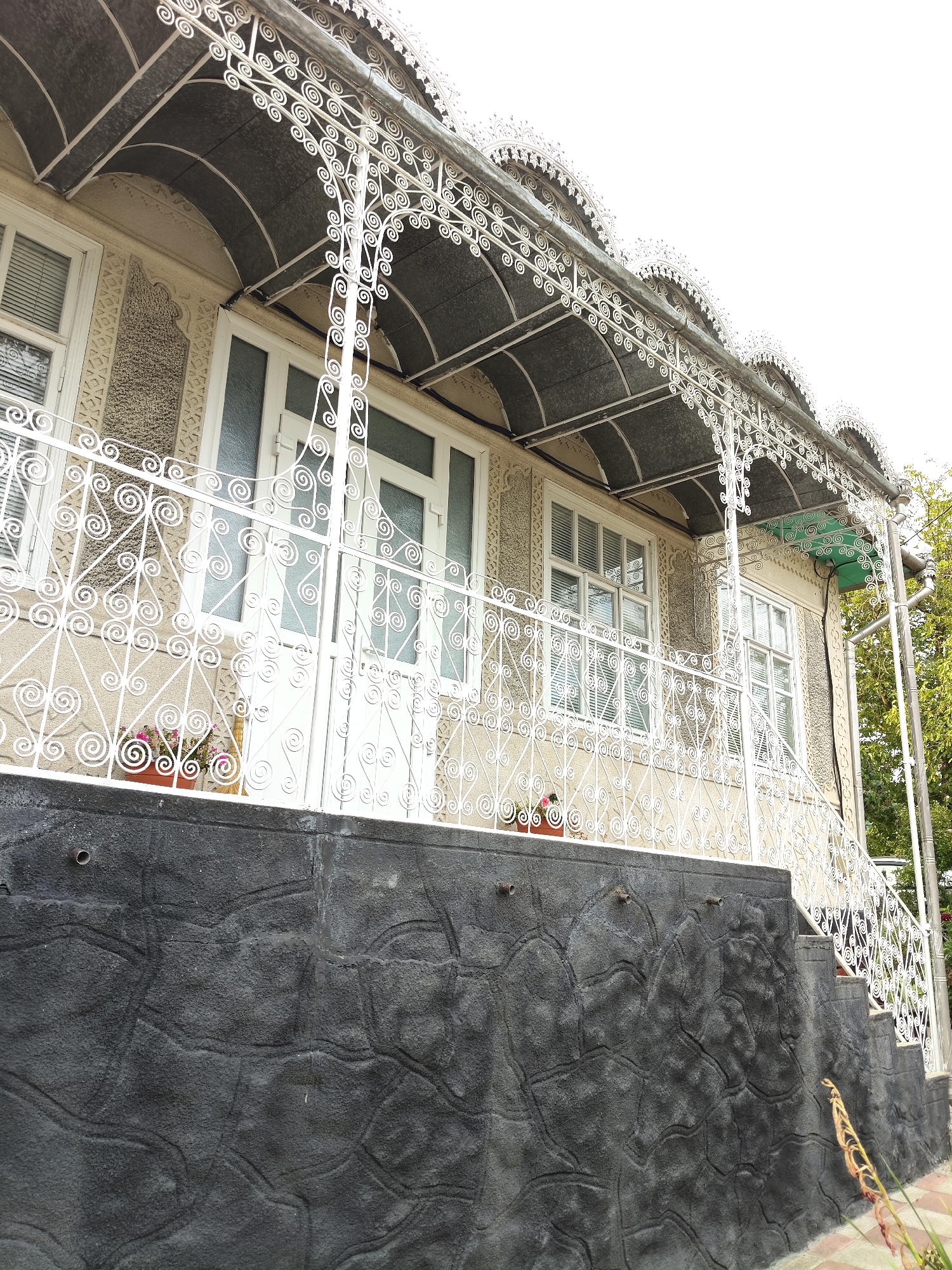 This is a medium-shot, outdoor, eye-level, wide-angle photograph of the facade of a two-story building. The building's exterior is painted a light beige color, with textured areas around the windows and doors, and decorative white metalwork on the porch and balcony railings. The porch has a dark, arched roof supported by ornate white metal pillars. The windows are white-framed with horizontal blinds, and there are two white doors on the ground floor, with a white railing in front of them. Small potted plants with pink flowers are visible on the balcony. The lower part of the building's foundation is a dark gray, textured concrete wall, with a rough, stone-like pattern. A set of concrete stairs with a white railing leads up to the right side of the building. The sky is overcast and bright white. Green trees are visible in the background to the right. The image was taken in the city of Gîrbova, Moldova.