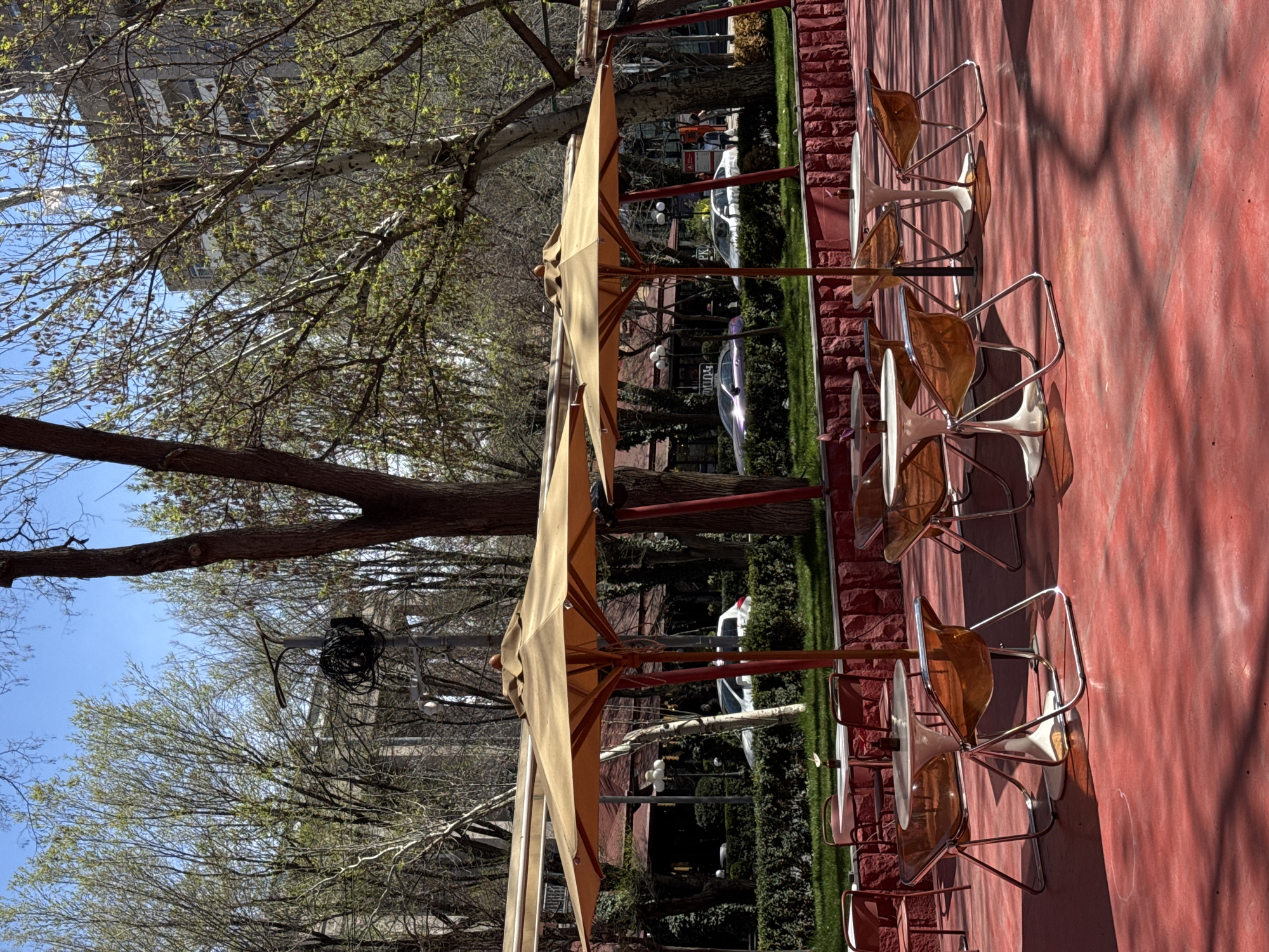 The image depicts an outdoor seating area of a cafe in Yerevan, Armenia. The setting features a bright red, textured concrete surface. Rows of metal-framed tables with cream-colored tops and transparent, amber-colored chairs are positioned under folded, light tan-colored umbrellas. The foreground is dominated by bare tree branches against a bright blue sky. In the background, the scene includes lush greenery, and cars parked along the road. Buildings are visible further back. The general atmosphere suggests early spring.