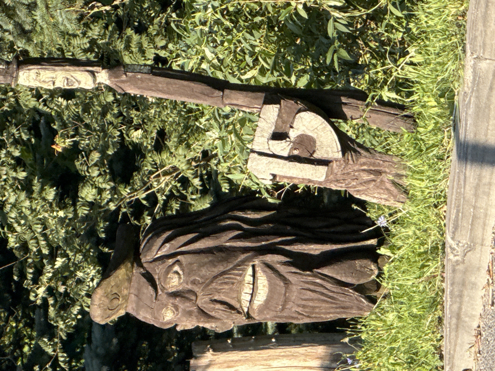 The image shows two wooden carvings set in an outdoor environment with green foliage in the background. The carving on the left is a bust of a face with exaggerated features, including wide eyes, a large nose, and an open mouth with visible teeth. It appears to be wearing a hat. To the right of this carving is a taller, more slender wooden figure that resembles a person. This figure has a carved face with an open mouth and is wearing what appears to be a hat and coat. Attached to this figure is a wooden sign with the number "15" carved into it. The carvings are weathered and appear to be made from tree trunks. In the foreground, there is green grass and a concrete curb. The background consists of dense green leaves and branches. The scene appears to be located in Galinė, Lithuania.