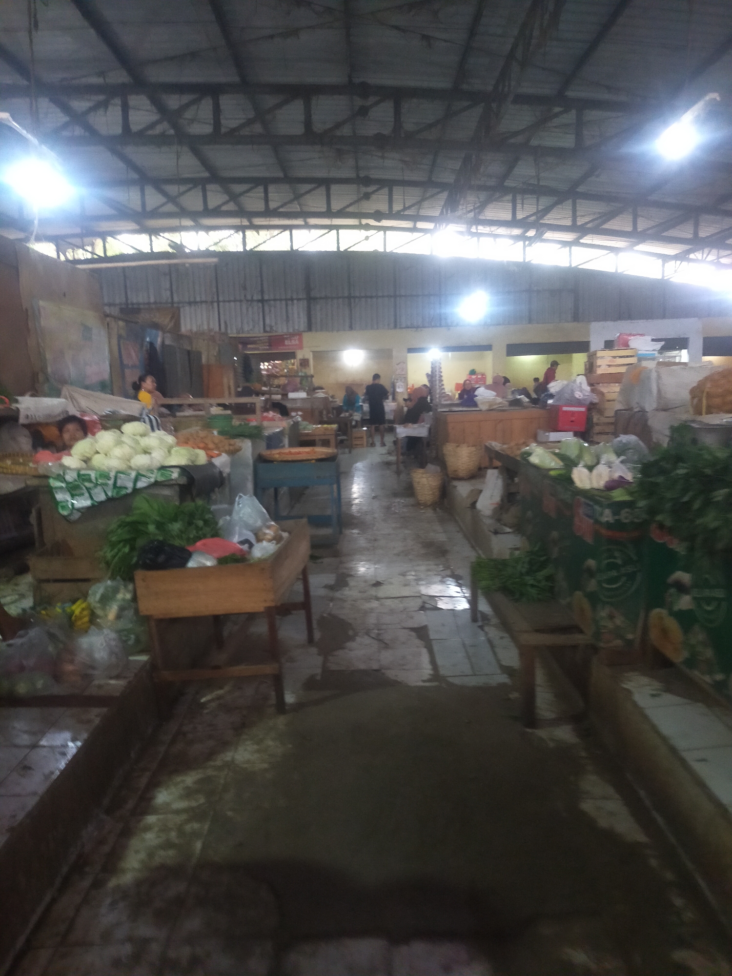 This is a slightly angled, low-light, full-shot image of the interior of a market stall, likely in Trayeman, Indonesia. The roof structure, composed of corrugated metal and exposed beams, is visible overhead, supported by a metal frame. Fluorescent lights illuminate the scene, creating bright spots and casting shadows.
The market appears to be selling produce, with various fruits and vegetables displayed on tables and in baskets. In the foreground, a wooden crate holds green vegetables and plastic bags of produce. To the left, a display table is covered with white cabbages, and a woman in a yellow top stands behind it. Further back, other stalls are visible, with people browsing and interacting. The floor is tiled and appears wet in some areas, reflecting the overhead lighting. The overall impression is that of a bustling, everyday market scene.