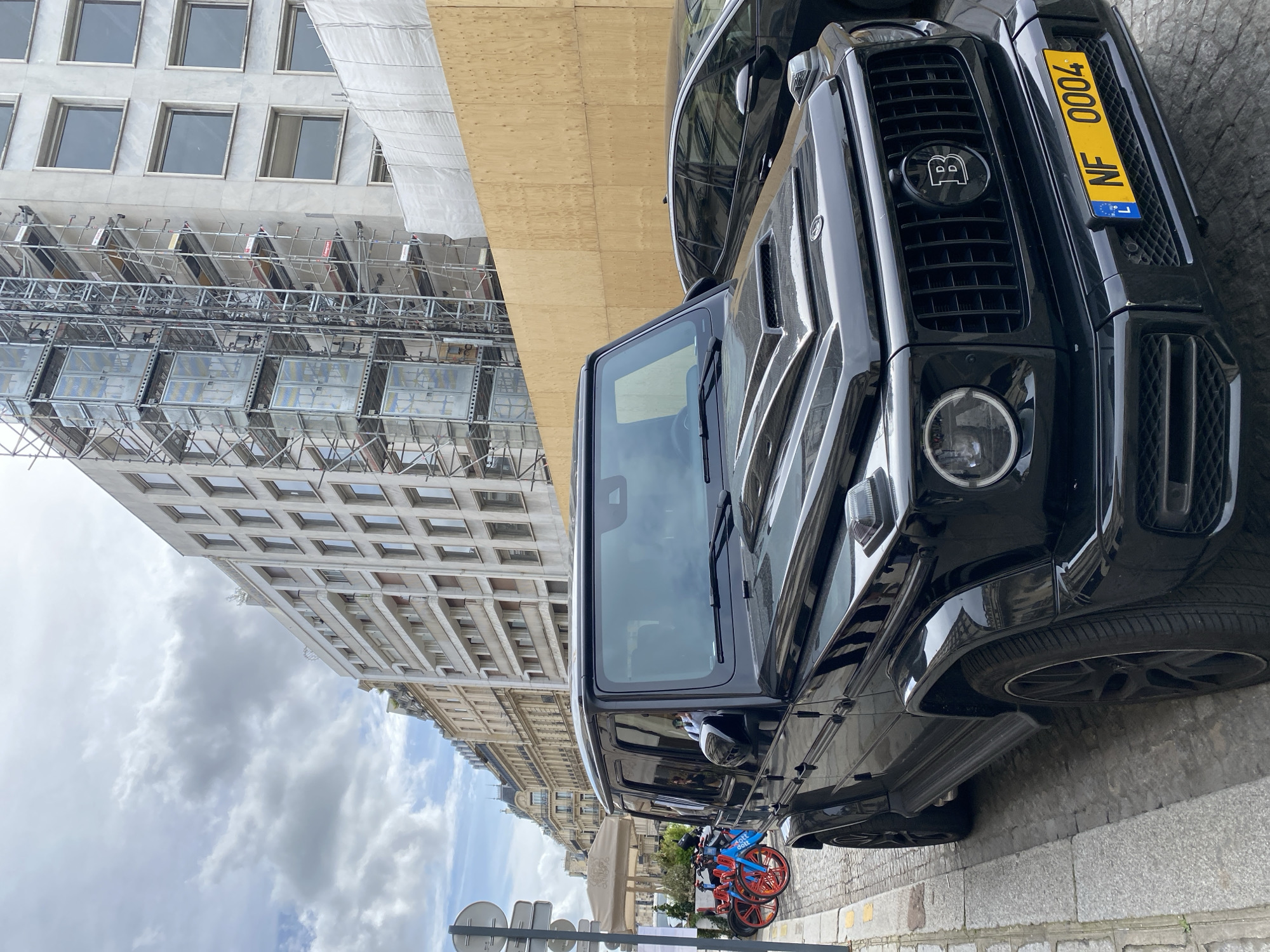 A black Brabus G-Wagon is parked on a cobblestone street, with its front end visible. The license plate is yellow with "NF 0004" printed on it, along with the flag of Luxembourg. The vehicle is positioned on the right side of the frame, adjacent to a sidewalk where blue and orange bicycles are parked. In the background, a tall building is under construction or renovation, covered in scaffolding. The sky is partly cloudy, with a mix of blue and grey. The scene is located in Paris, France, as evidenced by architectural details and street furniture in the background.
