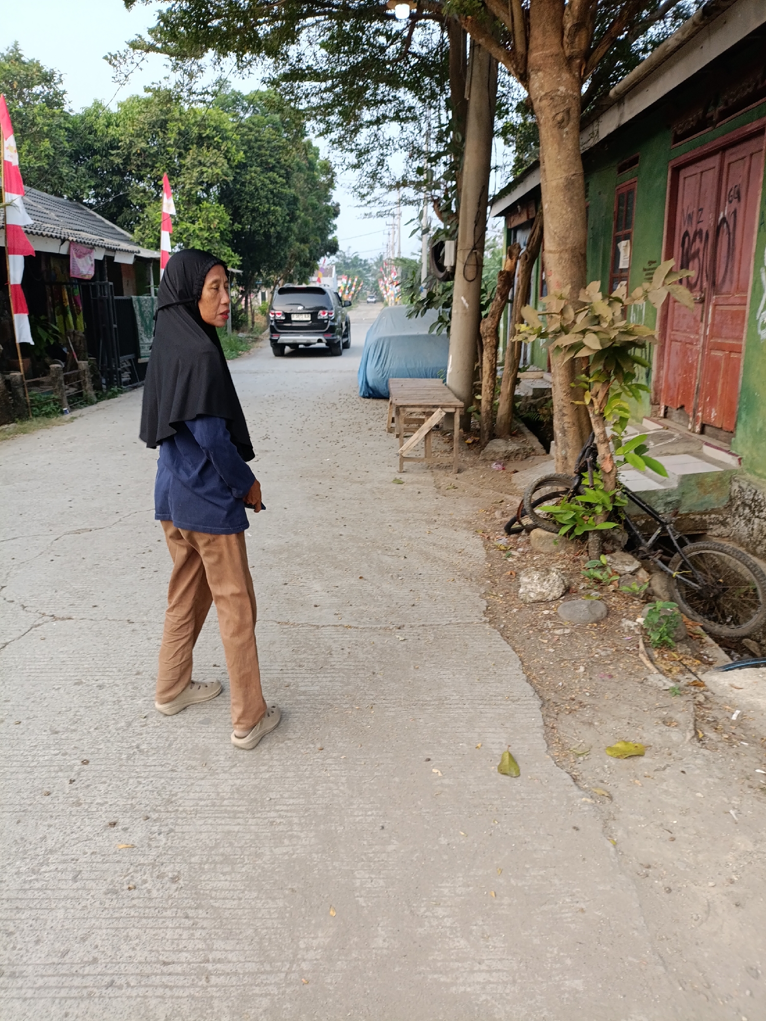An outdoor, medium-shot, eye-level, full-body photograph captures a woman walking on a concrete road in a village setting in Cariu, Indonesia. The woman, facing away from the camera and looking over her left shoulder, wears a black hijab, a dark blue long-sleeved shirt, and brown pants. She is also wearing light-colored shoes. The road is paved with concrete and appears slightly worn. To the woman's left, a black SUV is parked on the side of the road, and beyond that, a few houses with red and white flags are visible. To the woman's right, there is a green building with red doors, and a bicycle is leaning against a tree in front of the building. Trees and foliage are present throughout the scene, suggesting a rural or semi-rural environment. The sky is clear and overcast.