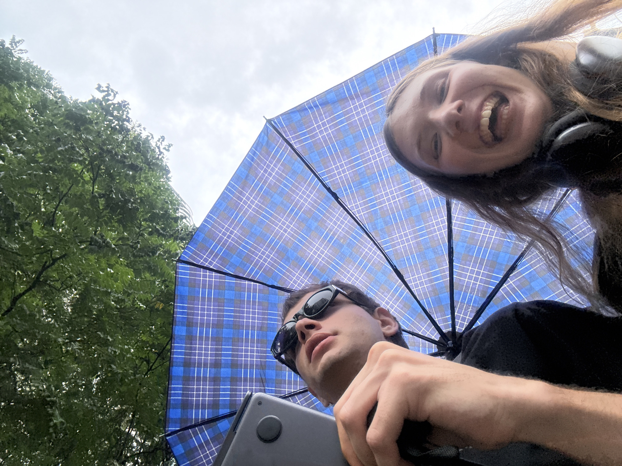 An ultra low-angle, upward-facing shot captures two individuals from below, positioned under an open blue plaid umbrella. The female, situated in the upper right, has long brown hair and a wide, joyful smile, revealing her teeth. She appears to be wearing headphones or an earbud on her left ear. To her left, in the lower frame, a male with dark sunglasses and a dark shirt looks slightly upward with a neutral expression. He holds a gray device, possibly a laptop or tablet, in his left hand. The blue umbrella, featuring a plaid pattern of varying blue and white lines, dominates the central part of the sky, providing a canopy over the subjects. To the far left, lush green tree foliage is visible against an overcast, light gray sky. The scene suggests an outdoor setting, possibly a park or green area, on a cloudy day, with the umbrella serving as protection from sun or light rain. There is no visible text in the image. This scene takes place in Jing'an District, China.