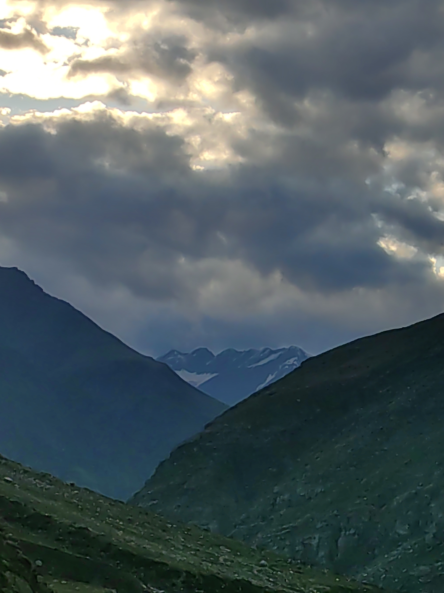 The image depicts a mountainous landscape under a dramatic sky filled with both dark, heavy clouds and bright, sunlit patches. The foreground features the steep, rocky slopes of mountains covered in sparse vegetation. Two prominent mountain ranges converge in the center of the frame, with the left range appearing darker and more shadowed, while the right range is bathed in a greener, more textured light. In the distance, further mountain peaks are visible, some with patches of snow. The sky is characterized by a gradient of dark blues and grays in the lower half, transitioning to brighter, white and yellow hues where the sun is breaking through the clouds. There are no people or man-made structures visible in the image, suggesting a remote, natural environment. The scene is consistent with the mountainous terrain found in regions like Damphug, India, known for its rugged topography and atmospheric skies.