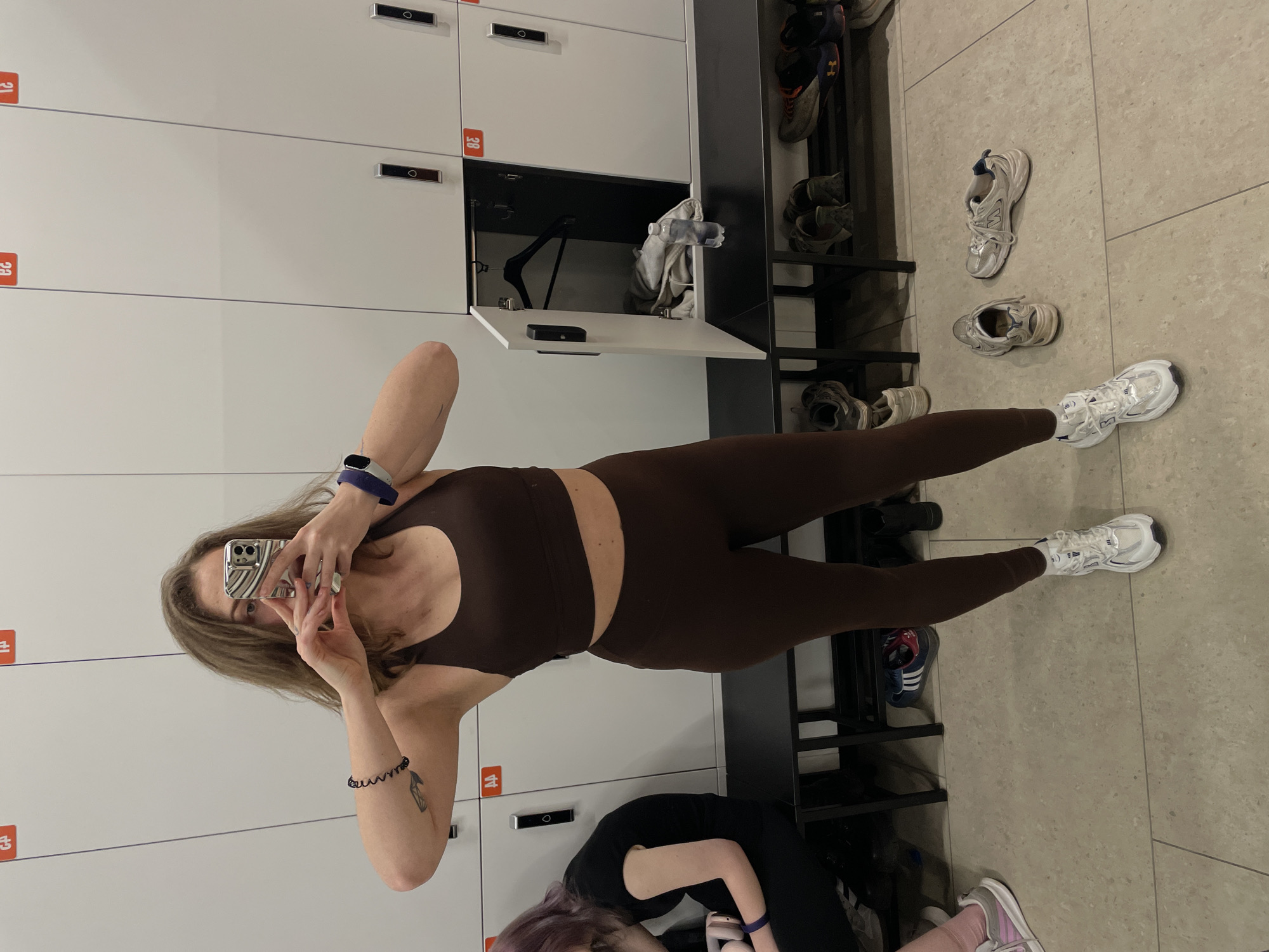 A woman in a brown sports bra and leggings is taking a selfie in front of a bank of white lockers with orange numbers. The lockers appear to be in a gym or changing room, with a shoe rack containing various pairs of shoes nearby. Another person, possibly sitting, is partially visible in the lower part of the image. The exact location in Moscow, Russia is not evident from the image.
