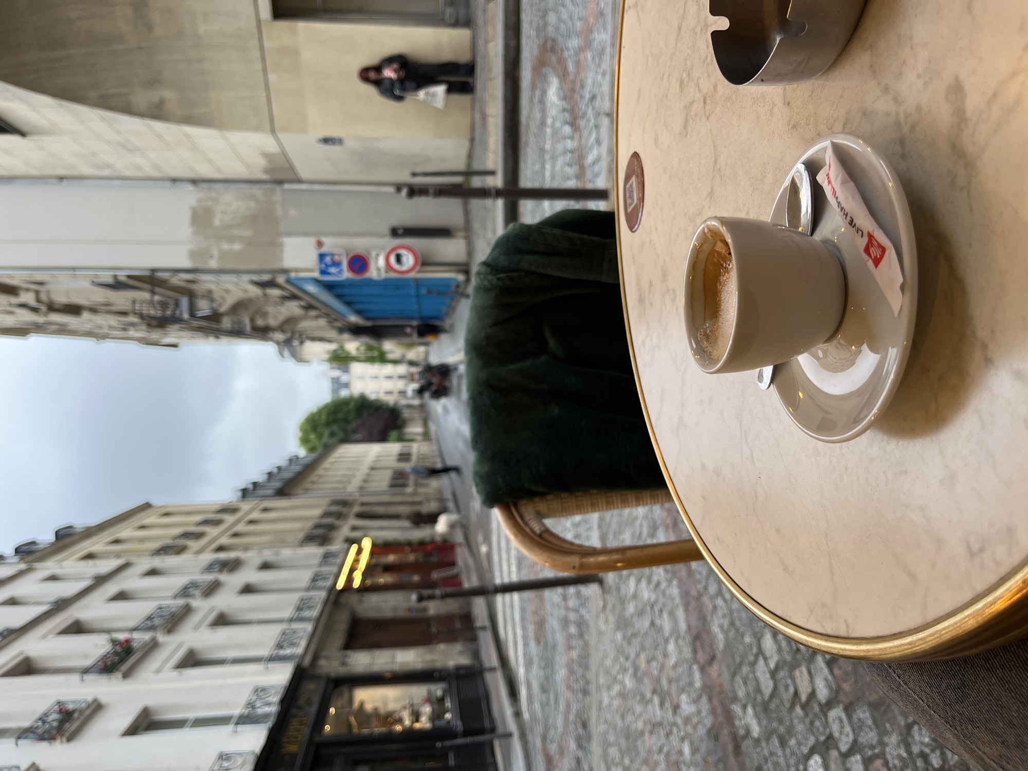 The image depicts an outdoor Parisian cafe scene on a rainy day. A cup of coffee and a sugar packet sit on a round table with a marble-like top and gold trim. An ashtray is also on the table. A dark green chair is partially visible behind the table.
In the background, the characteristic architecture of Paris is evident, including buildings with multiple stories, balconies, and intricate window designs. A street sign with regulatory symbols is visible along the road. People are walking on the wet cobblestone street. The sky is overcast and grey. The location is a typical Parisian street, lined with buildings and shops.