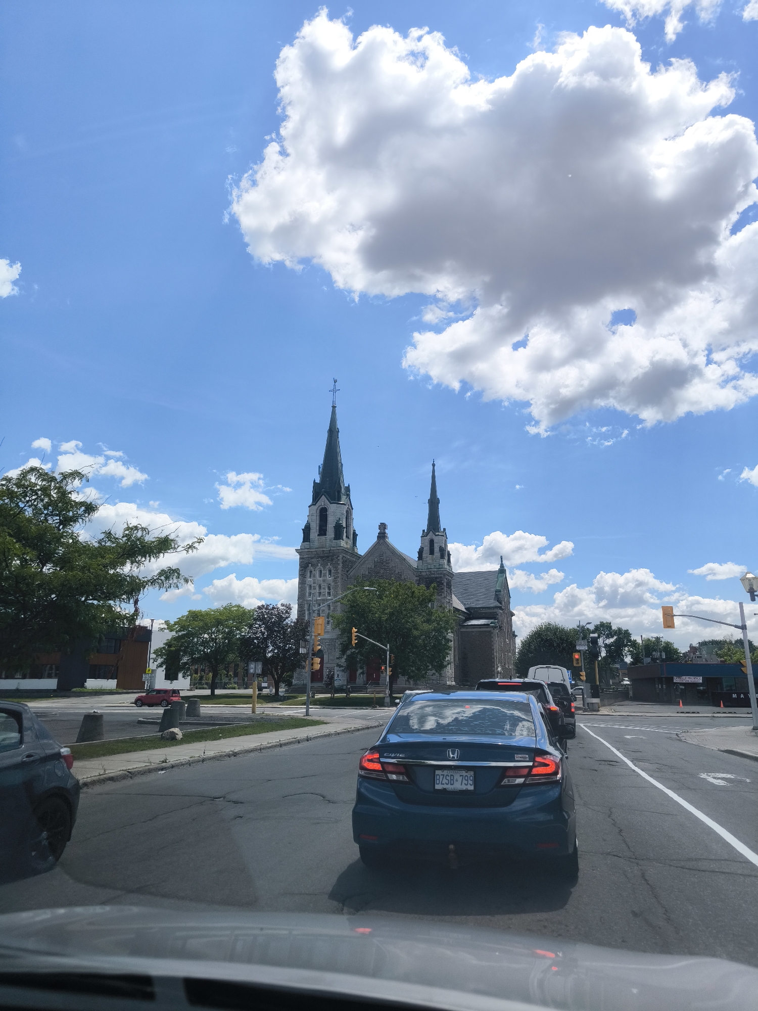 A wide-angle, eye-level shot captures a sunny day in Hawkesbury, Canada, dominated by a majestic stone church with a tall, green-spired steeple. The church is situated on the right side of the frame, its intricate architecture partially obscured by lush green trees. In the foreground, traffic moves along a paved road, with a blue Honda Civic in the center lane and a dark-colored car to its left. The road displays clear lane markings and some surface wear. Beyond the church, a few commercial buildings and traffic lights are visible, suggesting an urban setting. The sky above is a vibrant blue, dotted with fluffy white clouds.