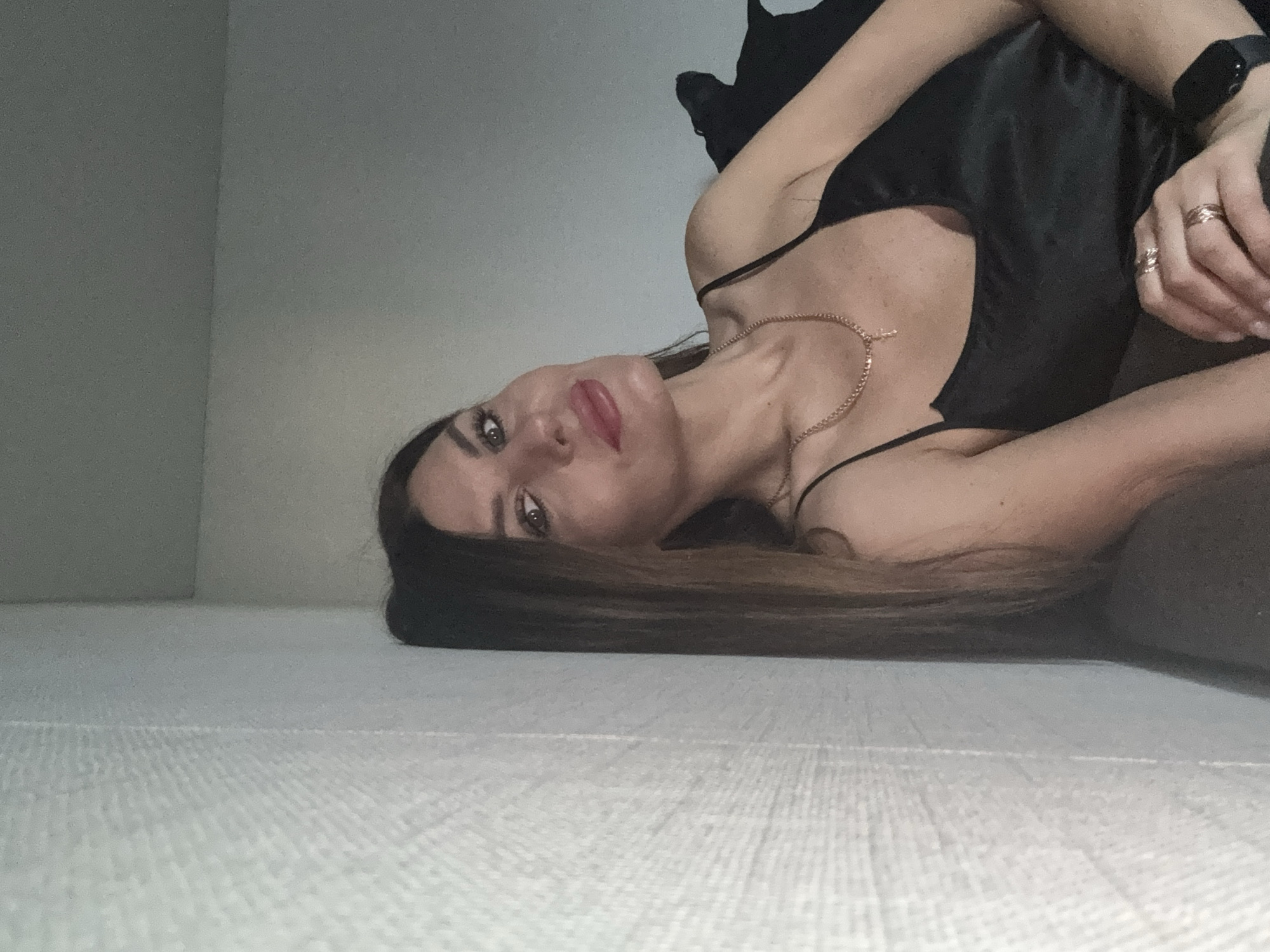 The image shows a woman lying on a light-colored textured surface, possibly a bed or carpet. She is wearing a black satin camisole and a gold necklace with a cross pendant. She has long brown hair, red lipstick, and is wearing rings on her fingers. A smartwatch is visible on her wrist. The background consists of a plain, light-colored wall. There is no specific information in the image to indicate the location as Chaikovsky, Russia.