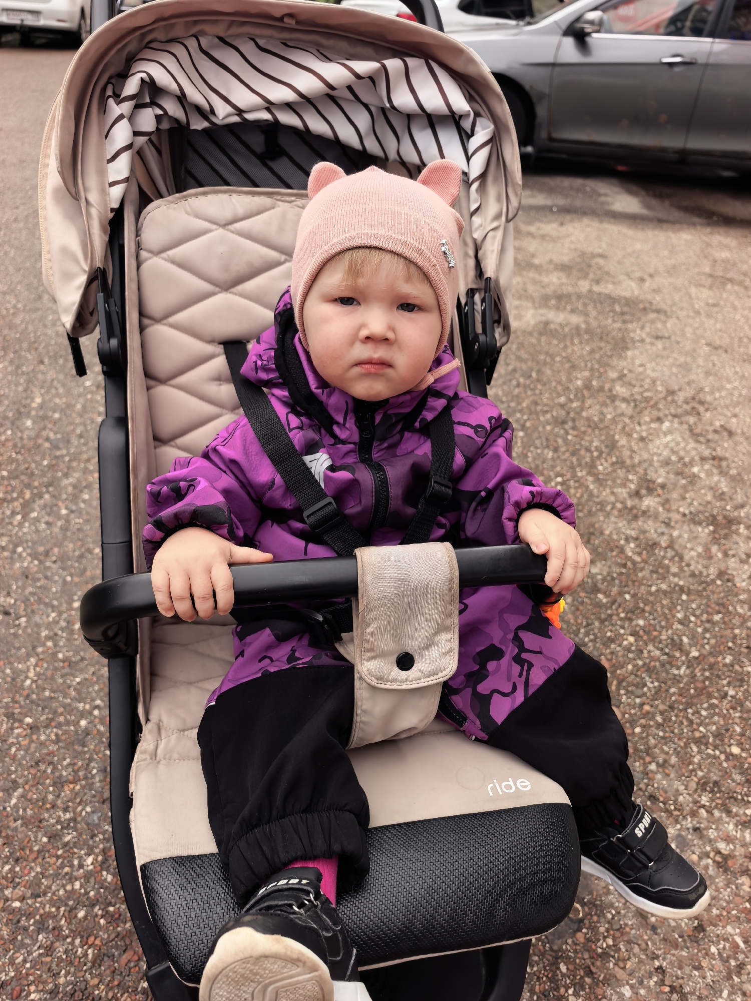 A young child with blonde hair and blue eyes, wearing a pink hat with bear ears and a purple camouflage jacket with black pants and pink socks, sits in a beige stroller. The child's hands are resting on the stroller's black safety bar. A beige pouch with a black button is attached to the bar. The stroller has a striped canopy. The child is looking at the camera with a neutral expression. The stroller is on a gravelly surface, possibly a sidewalk or parking lot, with cars visible in the background. The scene appears to be outdoors in an urban environment. The specific location of Sterlitamak, Russia cannot be determined from the visual information alone.