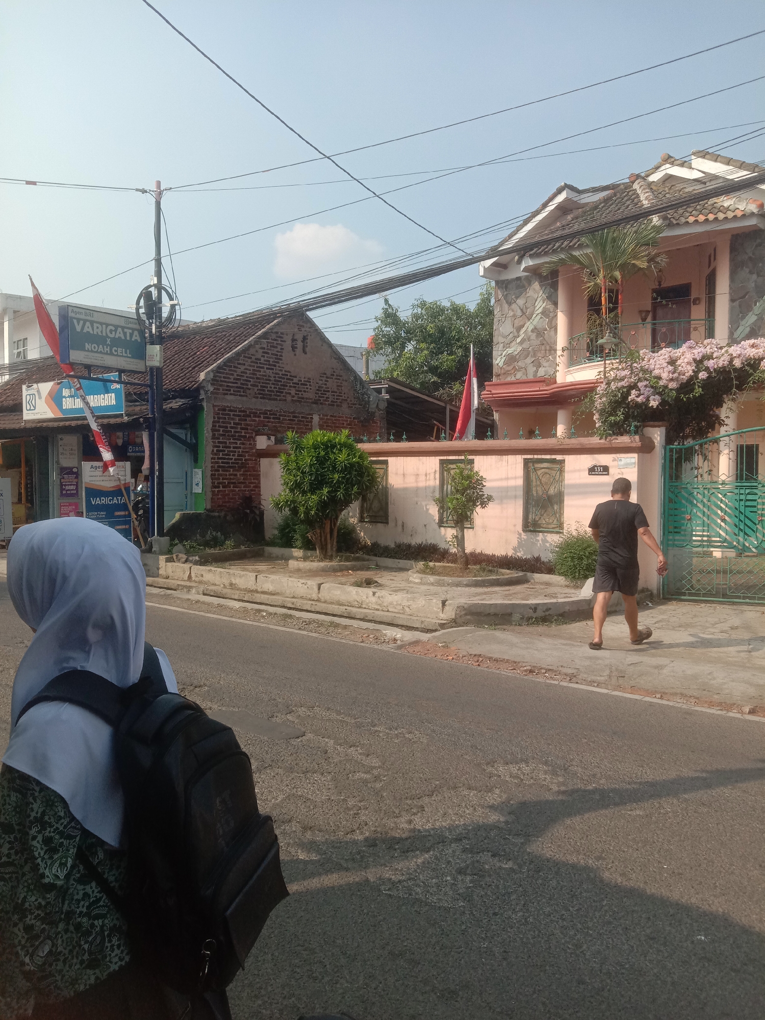 The image depicts a street scene in Cianjur, Indonesia, on a clear, sunny day. In the foreground, a person wearing a white hijab and a dark backpack is seen from behind, facing away from the camera and towards the street. A man in a black t-shirt and shorts walks on the sidewalk to the right of the street, looking away from the camera. The street is paved with asphalt and appears to be in good condition.
On the left side of the street, there is a small building with a sign that reads "Agen BRI VARIGATA X NOAH CELL". Next to it, another sign is visible with text including "Agen BRILink VARIGATA". A red and white flag is mounted on a pole in front of this building. Adjacent to this is a brick building with a tiled roof.
To the right of the street, there is a house with a pinkish-orange facade, stone accents on the upper story, and a tiled roof. A balcony overlooks the street, and blooming pink flowers cascade down from above. The house has a green metal gate. The number "131" is visible on the wall near the gate. The sidewalk is paved with concrete. Trees and shrubbery are visible in front of the buildings and house. Electrical wires crisscross the sky above the scene.