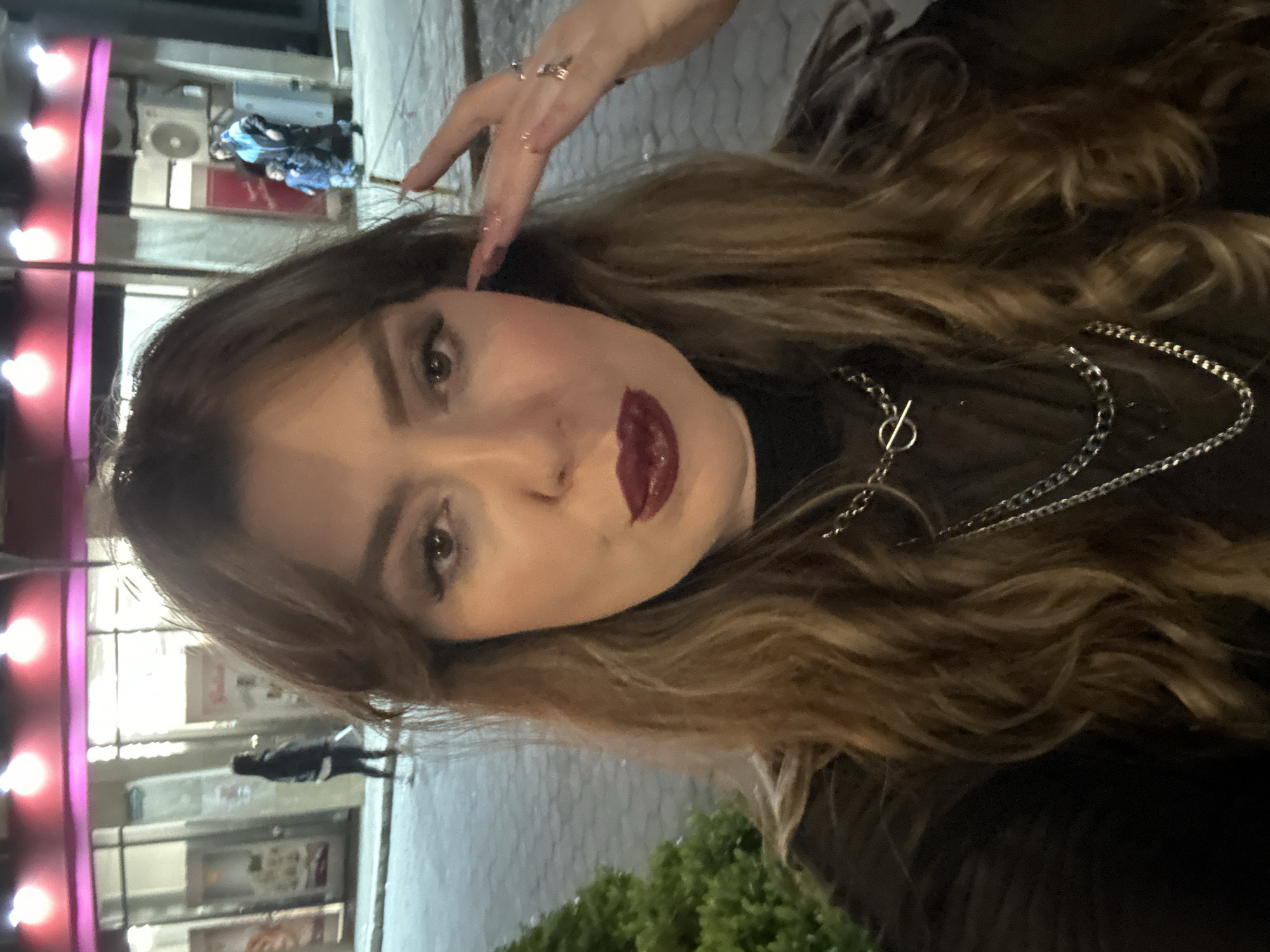 A woman is outdoors, possibly in Batumi, Georgia, based on the presence of buildings and streetlights typical of urban areas. The background includes a storefront with reflective windows, suggesting a commercial district. There are lights along the building, and signs are visible in the windows. The woman has dark hair with highlights, and she wears a silver chain necklace. Her lips are painted with dark lipstick. A hand is partially visible in the upper portion of the image, with nail polish. There are plants located near the bottom right of the photo.
