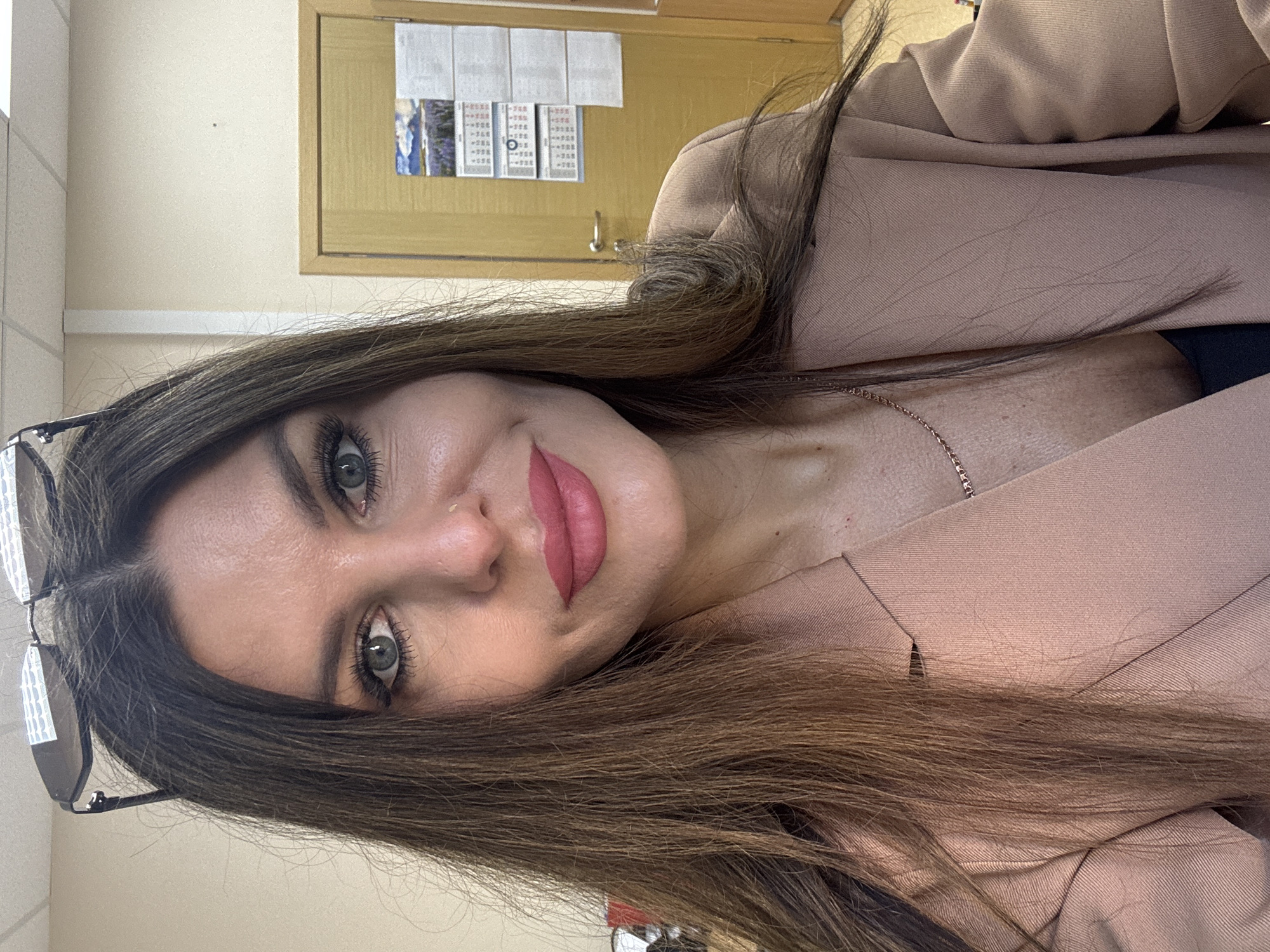 The image, taken in Chaikovsky, Russia, features a woman captured in a portrait-style shot, likely a selfie, with her head tilted. She is looking directly at the camera with a gentle, inviting smile. Her long, dark brown hair frames her face and extends across her shoulders and over a light beige blazer. She has striking green eyes, emphasized by prominent eyelashes and defined eyebrows. Her lips are adorned with pink lipstick. A small gold stud is visible on her right nostril, and a delicate gold-colored chain necklace can be seen around her neck, resting on her collarbone. Dark-rimmed glasses are perched on top of her head, partially visible on the left side of the frame. She wears a light beige blazer over a dark-colored top.
The background suggests an indoor setting, likely an office or institutional building. A light-colored wall, possibly cream or light beige, fills the upper left portion of the frame. A horizontal trim band runs along the wall. To the upper right, a light wooden door is visible, featuring a wooden-framed bulletin board with several papers pinned to it. Though the specific text is unreadable due to the angle and distance, patterns indicate lists or informational notices, possibly in Cyrillic script. A small blue and white image is also visible on one of the papers. A silver-colored hook is mounted on the wall below the bulletin board. The lighting suggests an indoor daytime scene.
