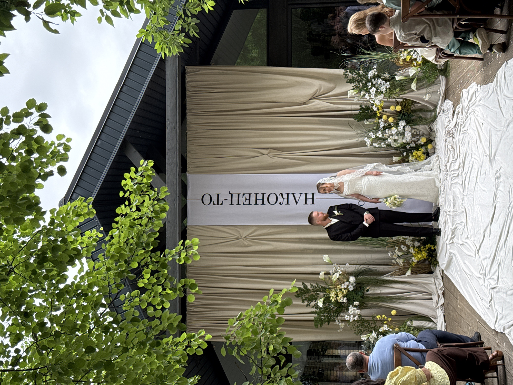 An outdoor wedding ceremony is captured in a wide shot, set in Perm, Russia. The bride and groom stand centrally on a raised wooden platform covered by a wrinkled white fabric aisle runner. The bride, positioned on the right, wears a white lace wedding gown with long sleeves and a flowing veil, holding a bouquet of white and yellow flowers. She gazes towards the groom with a smile. The groom, on the left, wears a black suit with a white shirt and a light-colored boutonnière, clasping his hands in front of him and smiling at the bride.
Behind the couple, a backdrop features beige curtains, with a tall white vertical banner inscribed with the Russian text "НАКОНЕЦ-ТО" (NAKONETS-TO), meaning "FINALLY!", centered behind them. Floral arrangements, predominantly white and yellow flowers with extensive green foliage, adorn both sides of the couple, flanking the banner and curtains. The ceremony takes place under a modern structure with a dark grey sloped roof and exposed beams, partially obscured by vibrant green tree foliage cascading from above. An overcast sky is visible through gaps in the leaves.
Guests are seated on wooden chairs on either side of the aisle. On the left, a man in a light blue shirt is seen from behind, and a person in a yellow top is partially visible. On the right, a young boy in a plaid shirt and green pants sits with his back to the camera, next to a partially visible woman with long blonde hair. Reflections of other guests and interior elements are visible in the glass panels behind the curtains on the right side of the venue.
