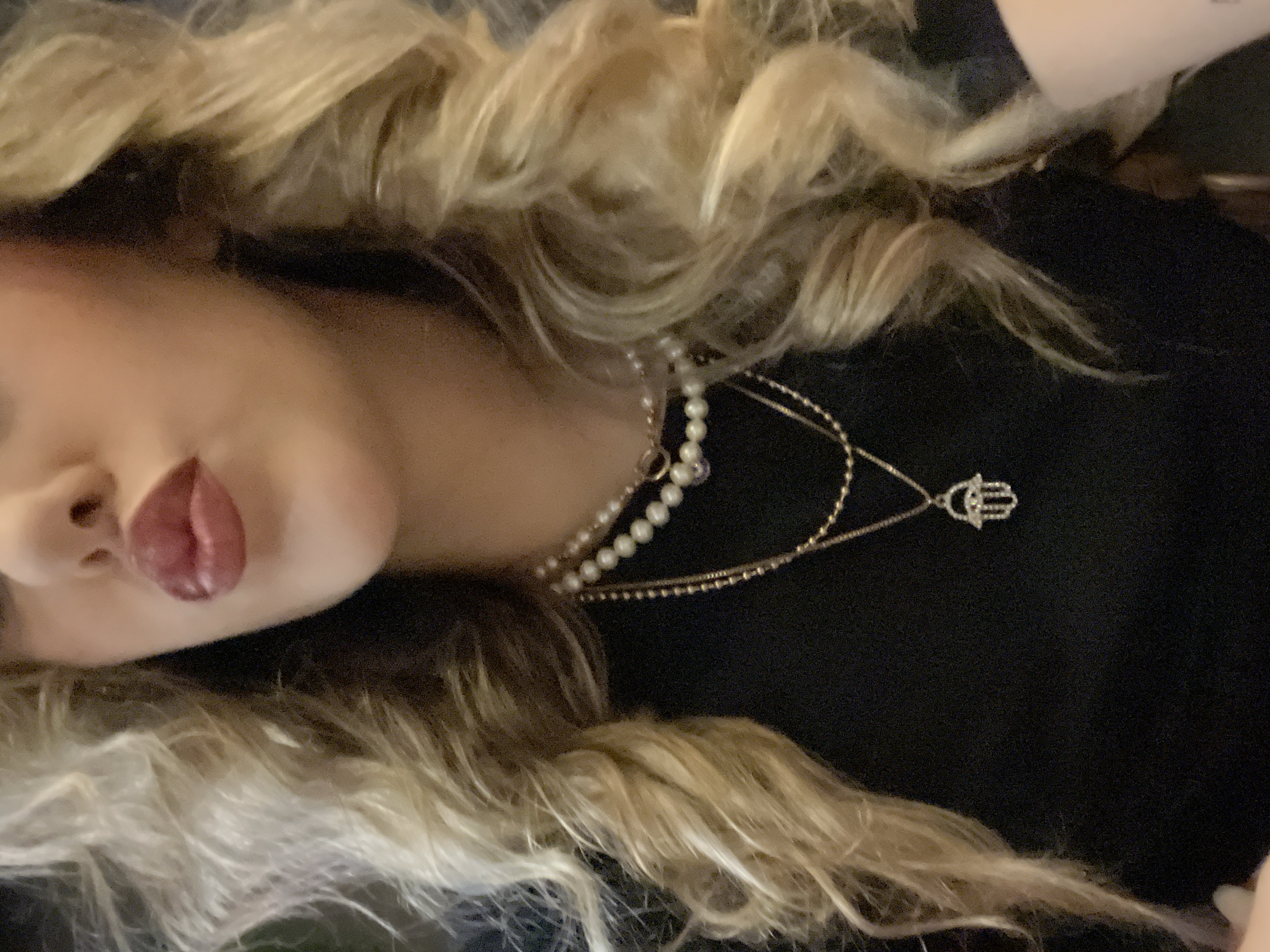 A close-up, slightly angled shot depicts a person's face and upper torso. The person has blonde, curly hair that frames the face and spills onto their black shirt. They wear a pearl necklace and a gold necklace featuring a Hamsa pendant. The person's lips are visibly pursed and colored with red lipstick. The background appears dark and indistinct. There is no evidence of the city of Sofia, Bulgaria or any other location.