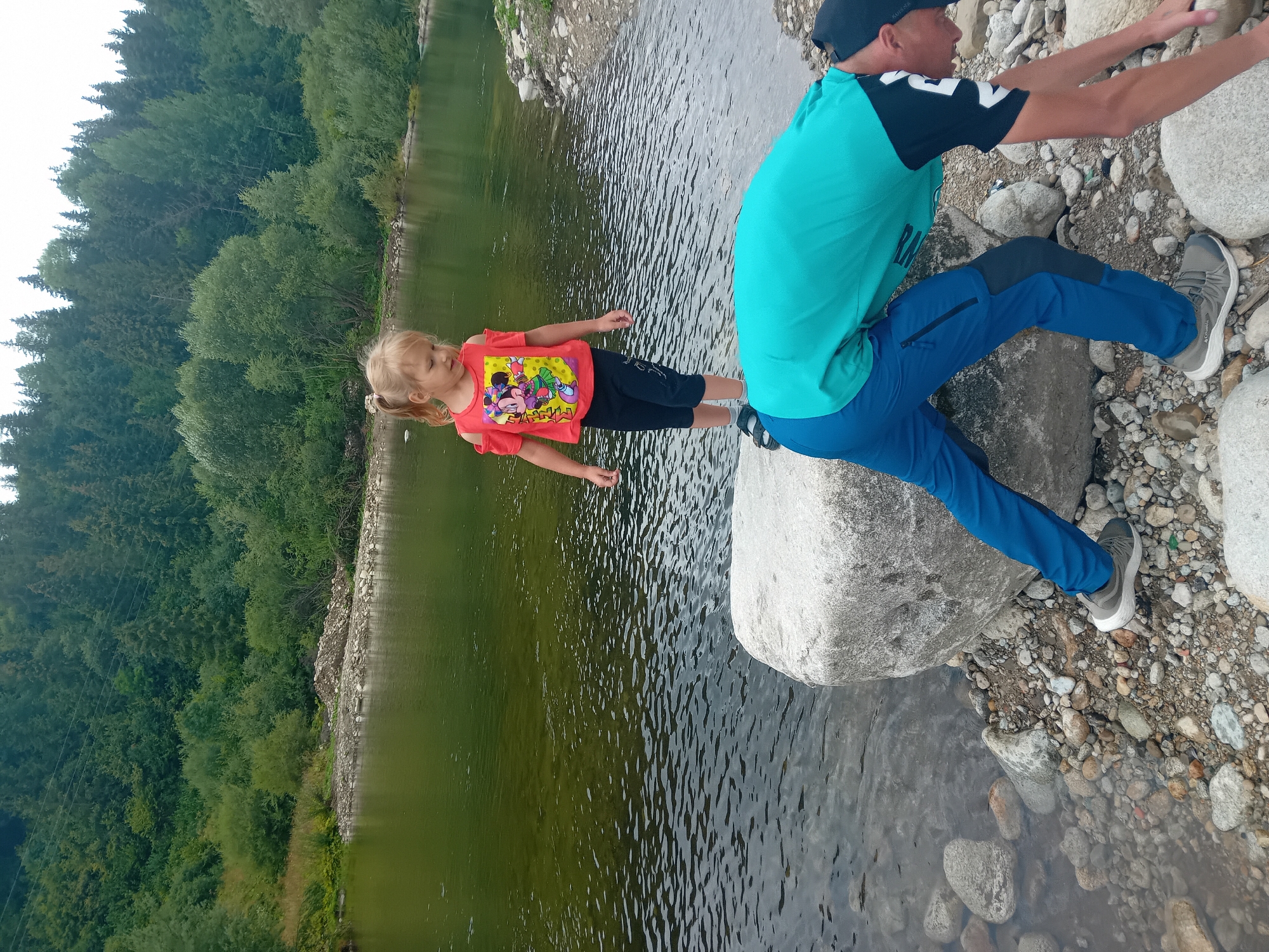 In Habovka, Slovakia, on a rocky riverbank, a young blonde girl with pigtails stands on a large rock overlooking a calm, green river. She is wearing a red t-shirt with a Minnie Mouse graphic and black capri pants. In the foreground, a person wearing a turquoise t-shirt and blue pants is bent over, appearing to reach for something near the river's edge. The background features a dense forest of green trees covering a hillside.