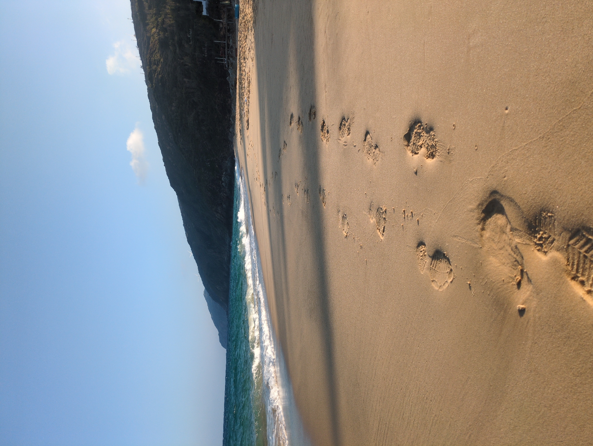 This image depicts a coastal scene on a sandy beach in East Quy Nhon Ward, Vietnam. The foreground shows light tan sand, prominently featuring a sequence of at least ten clear human footprints, possibly from a shoe, leading from the lower center towards the mid-ground and slightly to the right, receding into the distance. Long, dark shadows extend across the wet and dry sand from the left, indicating a low sun angle. To the left, turquoise-colored ocean water is visible with several white-capped waves breaking onto the shore. In the background, a dark, heavily vegetated mountainous or hilly coastline extends across the horizon, tapering towards the right. Faint structures, including what appear to be small buildings or foundations, are discernible at the base of the hill on the right. Higher on the distant hill, two white, vertical structures, consistent with wind turbine towers or similar infrastructure, are visible against the sky. The sky above is clear and light blue, with a small, wispy white cloud present above the right side of the hills. No individuals are visible in the frame, though the footprints indicate recent passage.