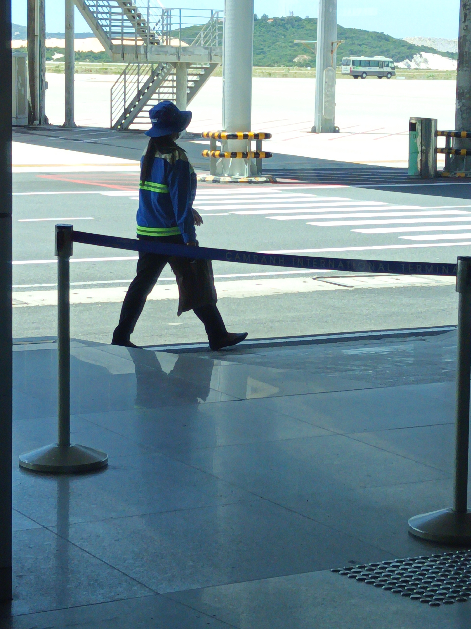 The image shows a view of Cam Ranh International Terminal in Bắc Cam Ranh, Vietnam. A person wearing a blue hat and a blue jacket with yellow stripes is walking. The person is wearing dark pants and dark shoes. A blue barrier tape stretches across the foreground. In the background, a bus is parked on the tarmac, and a set of stairs is visible. The ground appears to be tiled and reflective.