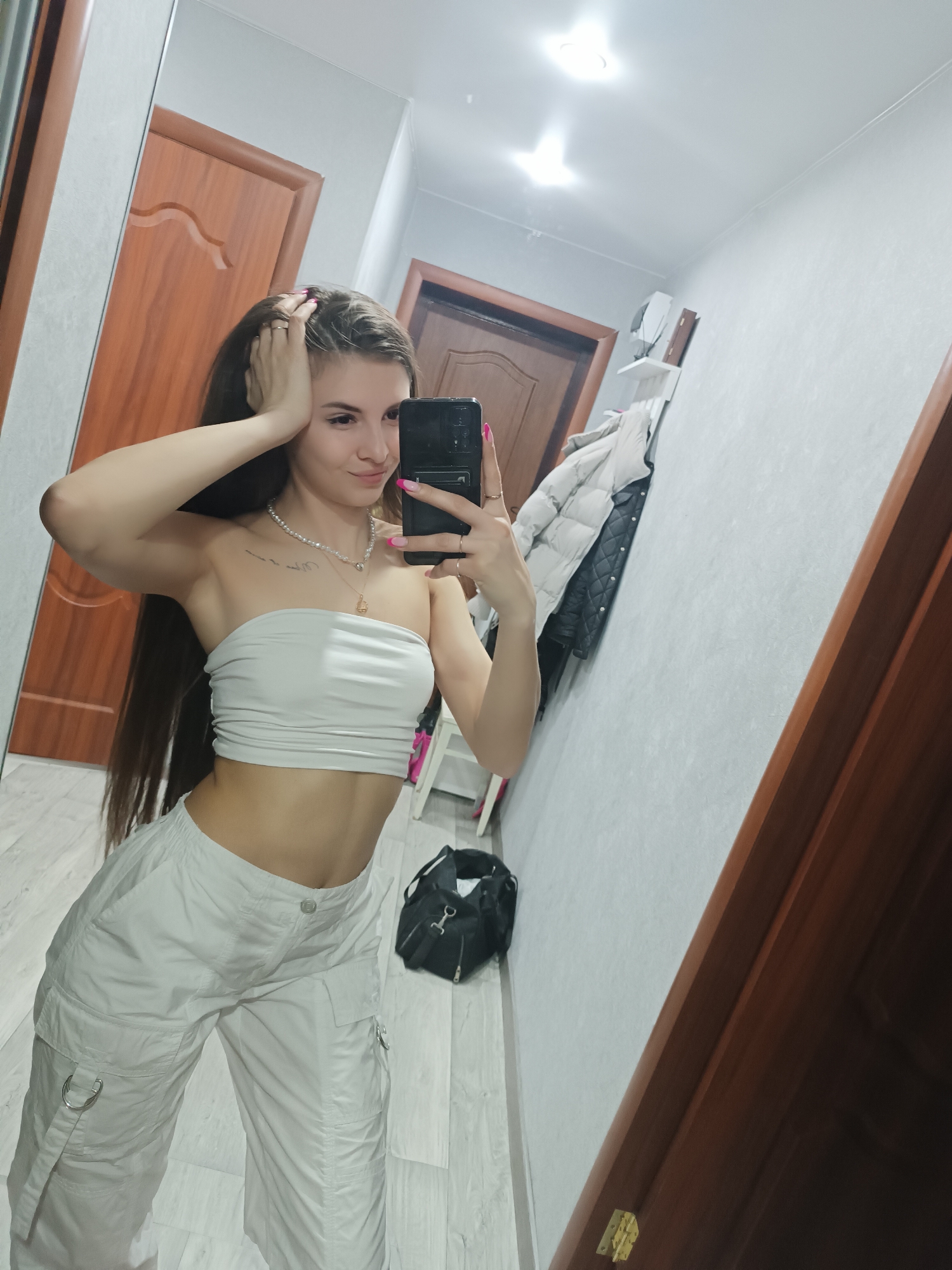 A woman takes a selfie in what appears to be a hallway. She is wearing a white tube top and white cargo pants. She has long brown hair and is wearing a necklace. She is holding her phone up to take the picture. Behind her, there is a closed wooden door and a white wall with a coat rack. A black bag is sitting on the floor. The location is possibly an apartment or home, but without further information, a precise location in Chaikovsky, Russia cannot be determined.