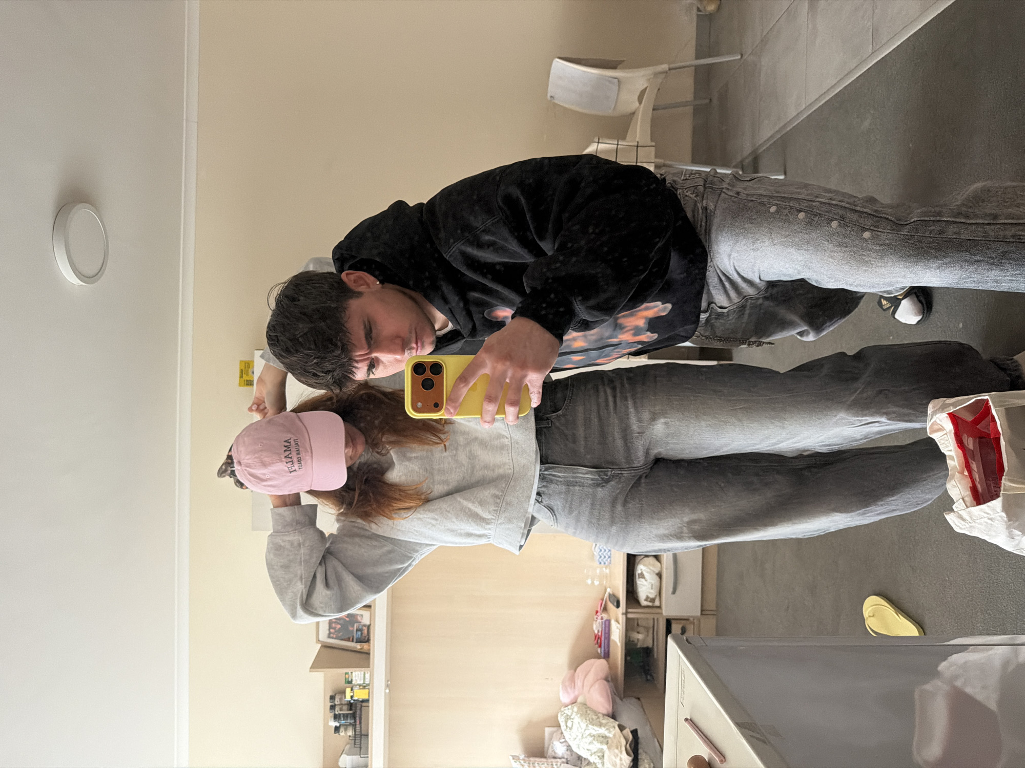 This media file captures a mirror selfie featuring two individuals inside what appears to be an apartment or room. The image is oriented sideways, with the ceiling to the left and the floor to the right.
**Subjects:**
The foreground features a young woman with reddish-brown hair, mostly covered by a pink baseball cap. She wears a light grey long-sleeved sweatshirt and faded grey jeans, and is holding a yellow smartphone (likely an iPhone) to take the picture, obscuring part of her face. Behind her stands a young man with short, dark hair. He is dressed in a black hoodie with a vibrant, possibly orange or red, graphic print on the front, and faded grey jeans. He looks downwards towards the phone, with a neutral expression. His visible foot wears a white sock and a black sandal.
**Objects:**
In the background, a light-colored chair is visible. To the right of the woman, shelves or cabinets are packed with various items like bottles, jars, and other household goods. A white, circular flush-mount ceiling light fixture is prominent on the ceiling. On the floor to the right, there's a white tote bag containing red items, and a yellow flip-flop. Below the woman's reflection, a pink plush object is visible among other small items on a surface.
**Scene and Setting:**
The setting is indoors, characterized by walls painted in contrasting light beige and white tones, suggesting an interior corner. The floor is a grey, possibly concrete or tiled, surface. The lighting is bright and even, indicative of indoor illumination.
**Activity:**
The two individuals are posing for a casual mirror selfie, with the woman actively holding the phone.
**Notable Details:**
The time of day is not discernible. No specific location cues within the image itself indicate Sofia, Bulgaria. The general ambiance is relaxed and informal.
**Visible Text:**
Text on the pink baseball cap is reflected and reads "AMALIJ ATTIYA" (mirrored).