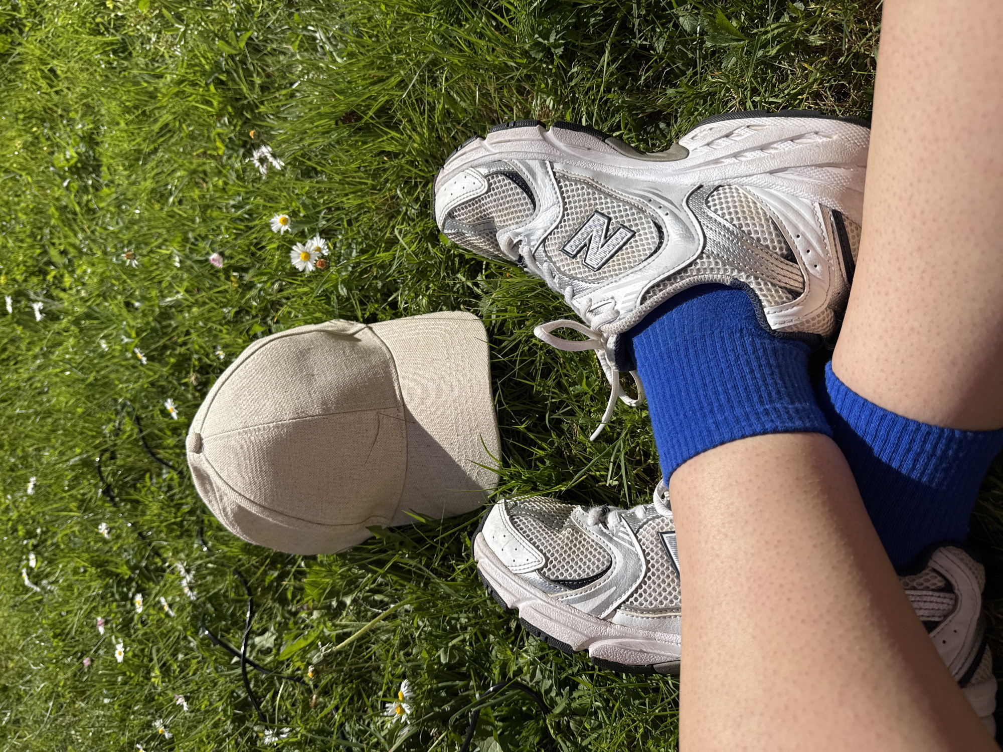 An overhead shot captures a person's lower legs and feet, along with a cap, resting on vibrant green grass dotted with small white daisies. The scene is bathed in bright, direct sunlight, suggesting a clear and sunny day, likely midday or early afternoon.
The person is wearing a pair of white and grey New Balance 530 sneakers, characterized by their mesh and synthetic leather construction and the prominent 'N' logo on the sides. Underneath the sneakers, bright royal blue crew socks are visible, creating a striking color contrast with the shoes and grass. The skin on the exposed lower legs appears to have a natural texture with small visible pores.
Next to the person's feet lies a light beige or cream-colored baseball cap, made from a woven fabric, possibly linen or cotton. Its brim is facing away from the viewer.
The setting is a natural grassy area, likely a park or lawn, in Great Yarmouth, United Kingdom. The scattered daisies and lush green provide a fresh, outdoor ambiance. The overall impression is one of relaxation and comfort, with the person enjoying a moment of rest outdoors in pleasant weather. No explicit activity beyond resting is depicted. There is no other visible text apart from the New Balance logo.