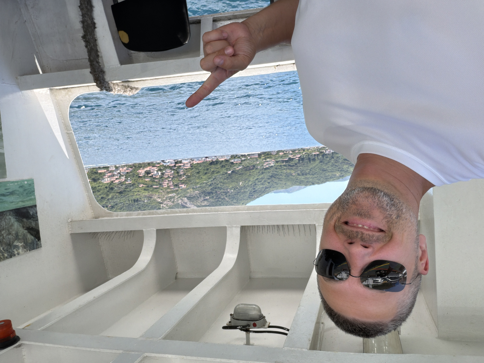 A selfie is taken by a man with short, graying hair and a stubbled beard, wearing sunglasses and a white t-shirt. He is on a boat, with the blue sea and a hillside town visible in the background. The town has many red-tiled roofs and is nestled against a green, tree-covered mountain. The boat's interior, with white beams and a light fixture, is also visible. The man is pointing his right index finger towards the water. The location is identified as Lepetane, Montenegro.