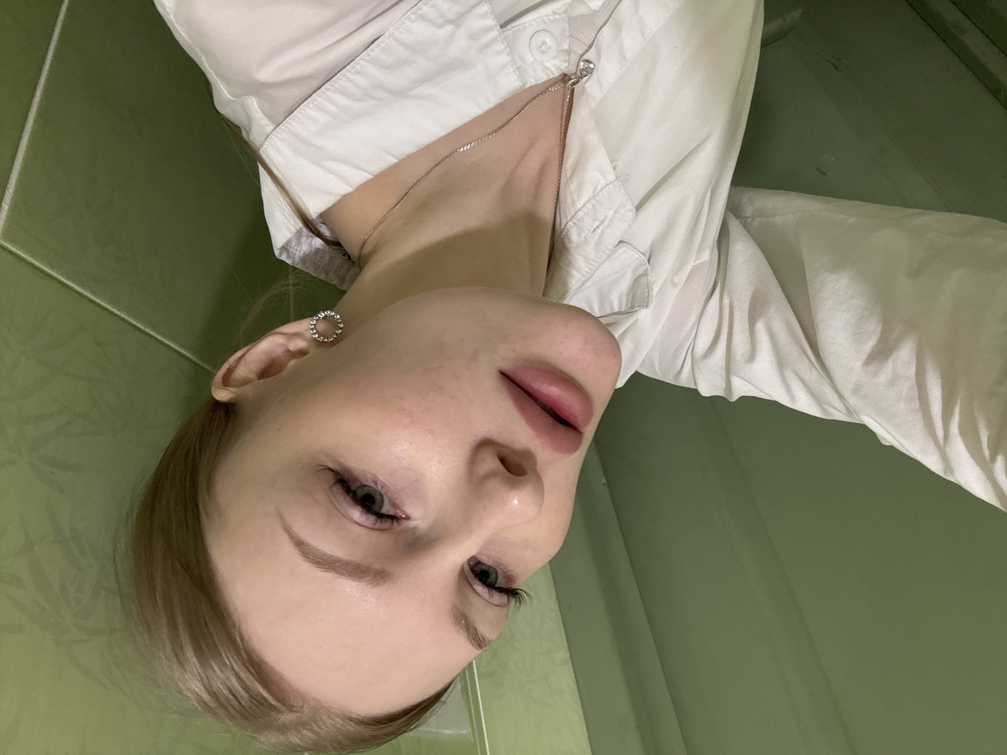 A close-up, slightly downward-angled shot features a woman with light skin and blonde hair partially visible in the lower left corner. She has green eyes, defined eyebrows, and plump lips. She wears an earring in her left ear and a silver necklace. She is wearing a white collared shirt. The background is a green wall, possibly tiled, suggesting a bathroom. There is no specific evidence in the image to place this scene in Perm, Russia, or any city.