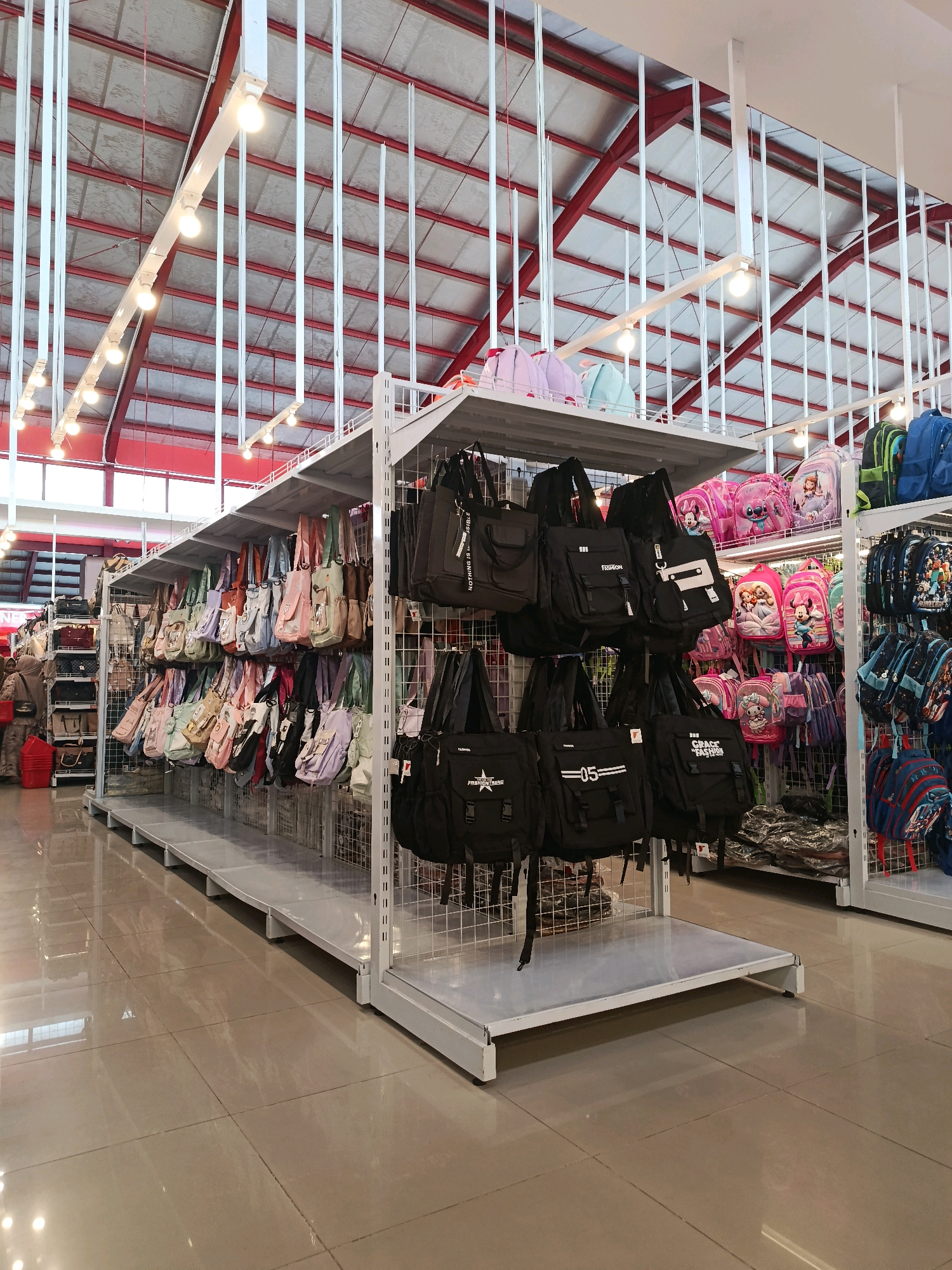 The image depicts the interior of a retail store, likely a shop selling bags and backpacks, located in Bulukumba, Indonesia. The store features multiple shelves displaying a variety of bags in different colors and styles. On the right side, there are racks filled with colorful backpacks, including some with animated characters, and on the left, a wider assortment of tote bags and shoulder bags. The store has a high ceiling with exposed red beams and a silver corrugated roof, illuminated by rows of white tube lights. The floor is a polished tiled surface, reflecting the overhead lighting and the merchandise. In the background, to the left, a person wearing a hijab is partially visible, browsing the merchandise, and a red plastic bucket is on the floor. The overall impression is that of a well-stocked retail environment.