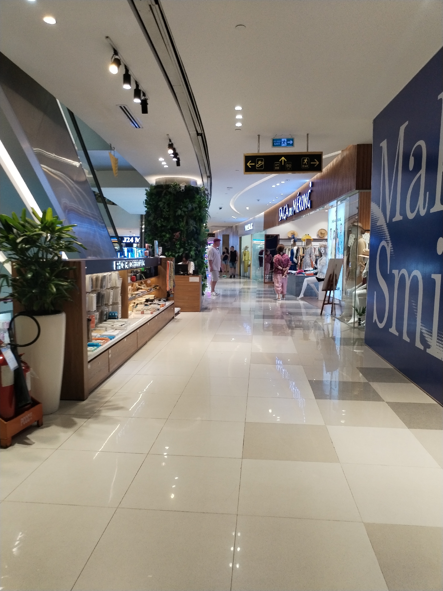 The image depicts an indoor shopping mall environment in Nha Trang Ward, Vietnam. The perspective is from a slightly low angle, looking down a wide, brightly lit corridor with a polished tiled floor that reflects the overhead lights.
On the left side of the corridor, there is a tall, white planter with a lush green plant. Next to it, a red fire extinguisher is visible on a mobile orange base. Further down the left, a display counter is set up, showcasing various items like phone cases and accessories. Behind the counter, a large vertical green wall of plants serves as a decorative element.
The right side of the corridor features several storefronts. One prominent storefront has the name "SAGA du MEKONG" displayed above it. The windows of these stores reveal mannequins dressed in clothing and displays of merchandise. A large blue sign with white lettering, partially visible on the far right, appears to be part of a store's branding.
Several individuals are present in the scene. A man in a white shirt and shorts stands near a storefront on the right. Further down the corridor, a woman in a patterned pink outfit is walking away from the camera. Other people can be seen in the distance, browsing in the shops.
Overhead, the ceiling is white, with a series of track lighting fixtures. Signage is visible on the ceiling, including directional arrows for exits and stairs. The overall impression is a modern, clean, and well-maintained shopping area.