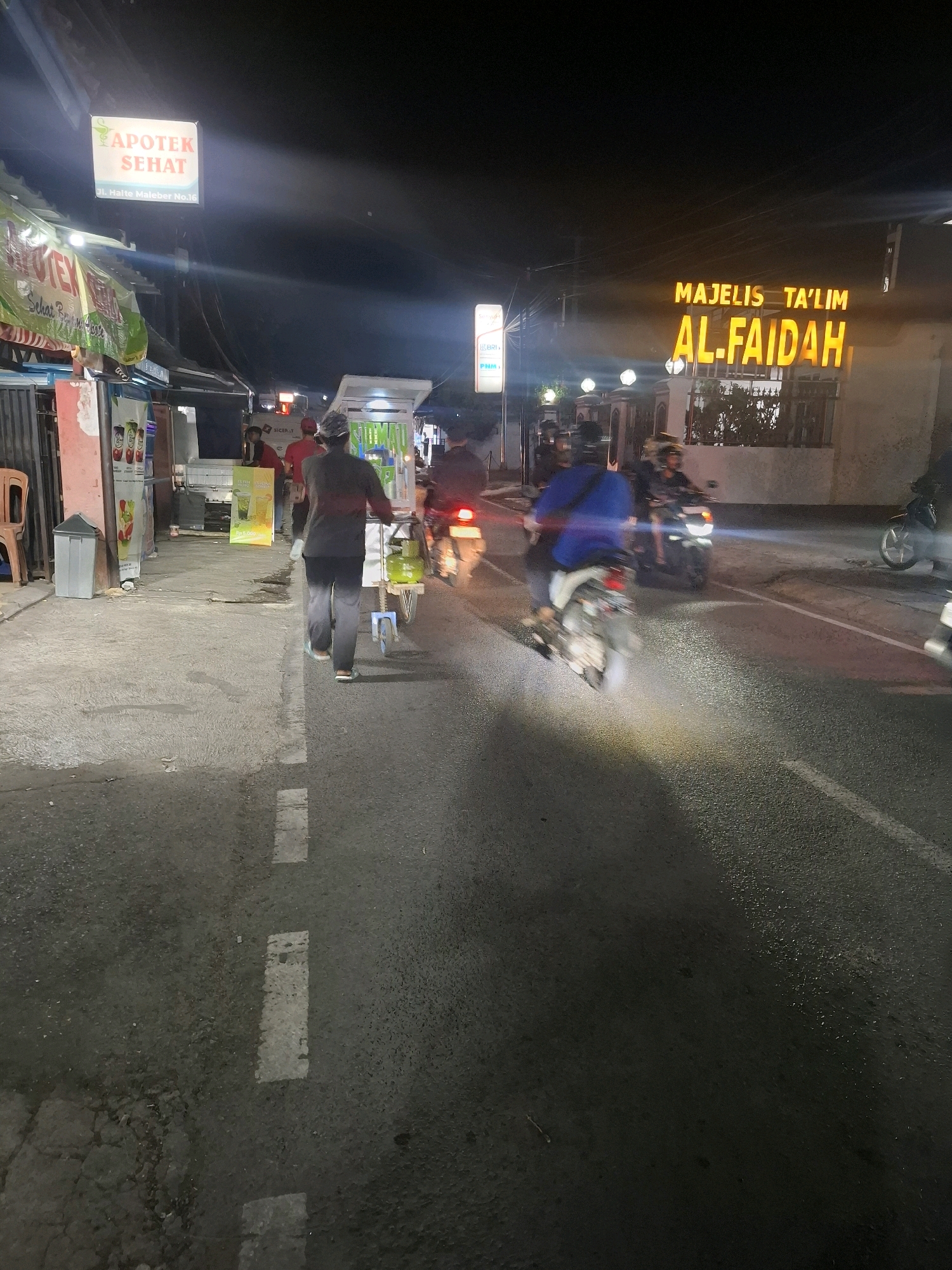 This image captures a nighttime scene on a street in Karangtengah, Indonesia. A prominent sign illuminated with orange light reads "MAJELIS TA'LIM AL-FAIDAH." To the left, another illuminated sign says "APOTEK SEHAT" with "Jl. Halte Maleber No.16" below it, suggesting a pharmacy. The street is paved with asphalt, marked by white dashed lines. Several motorcycles are visible, with some riders in motion, their headlights cutting through the darkness. One person is pushing a cart with a gas cylinder along the sidewalk. The environment appears to be commercial, with shops and stalls lining the street, some displaying colorful advertisements. The overall atmosphere is lively, illuminated by various artificial lights, indicating a bustling urban night.