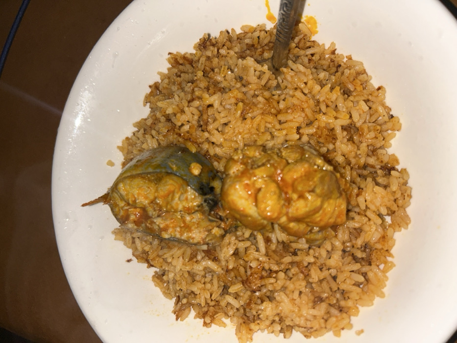This scene is situated in Egbe, Nigeria. The primary subject is a white, round, ceramic bowl containing a meal. The bowl's upper rim shows small water droplets. The meal consists of light brown and orange-tinted rice, uniformly mixed with reddish-brown spice particles. Two pieces of cooked fish are positioned on top of the rice. The upper fish piece displays sections of bluish-grey skin and flesh covered in an orange-red sauce, with a pointed bone or fin protruding upwards. The lower fish piece is fully coated in a similar orange-red sauce, revealing textured fish meat. A metallic eating utensil, appearing to be a spoon or fork, is partially visible on the right side of the bowl, embedded in the rice. The immediate background is dark and out of focus, consistent with an indoor setting under subdued lighting.