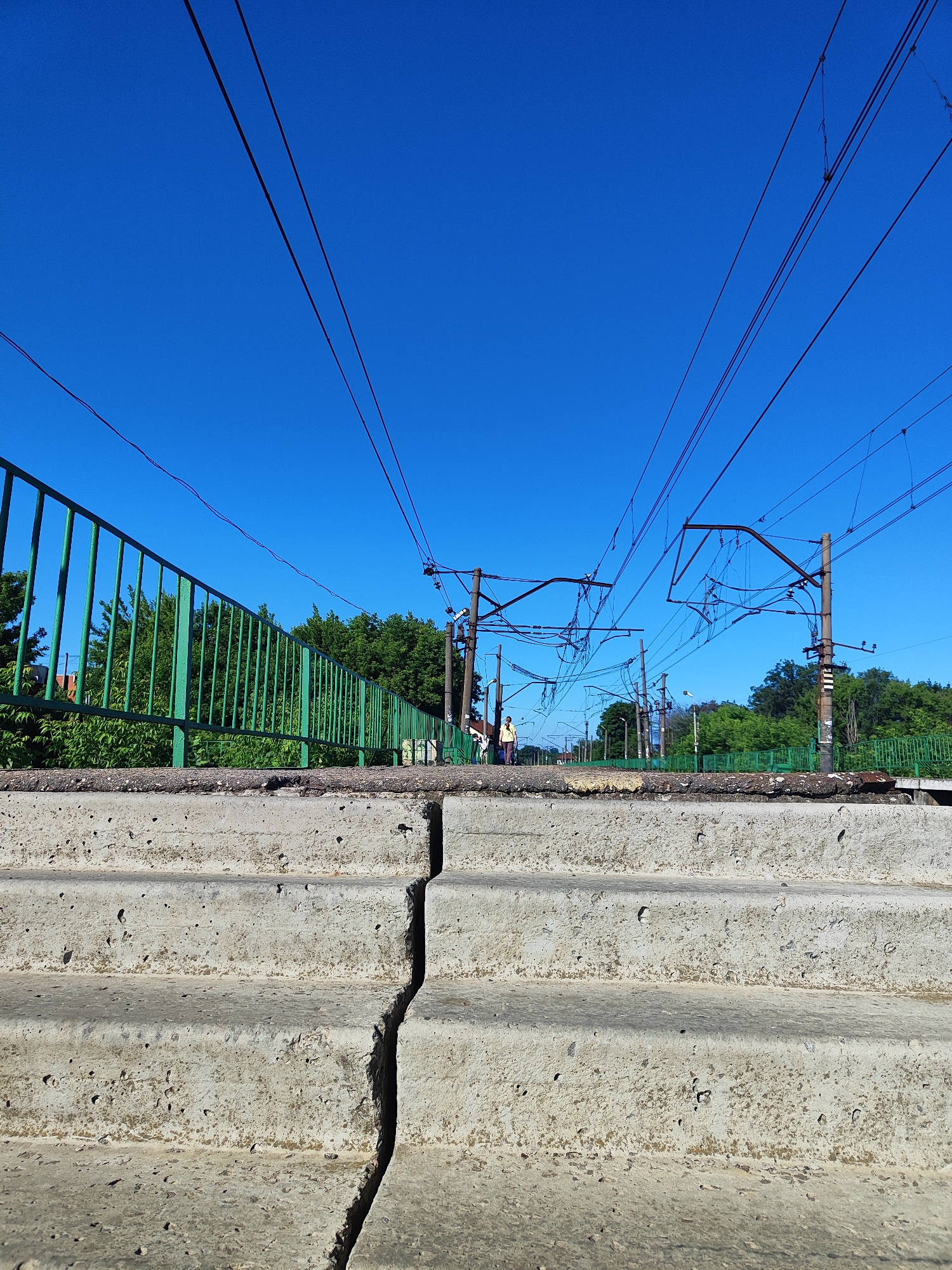 An eye-level, medium shot captures a section of outdoor railway infrastructure against a bright blue sky. In the foreground, a cracked concrete structure, possibly stairs or a platform edge, dominates the lower half of the frame. Its surface is rough and weathered, with visible damage and discoloration. Behind this concrete element, a green metal railing runs horizontally, separating it from a dense backdrop of green trees. Above the trees and railing, a complex network of overhead power lines and support poles stretches across the sky, indicating an active railway line. A lone figure, dressed in yellow and white, stands on what appears to be a railway platform in the distance, facing left. The overall scene is one of urban infrastructure bathed in bright sunlight.