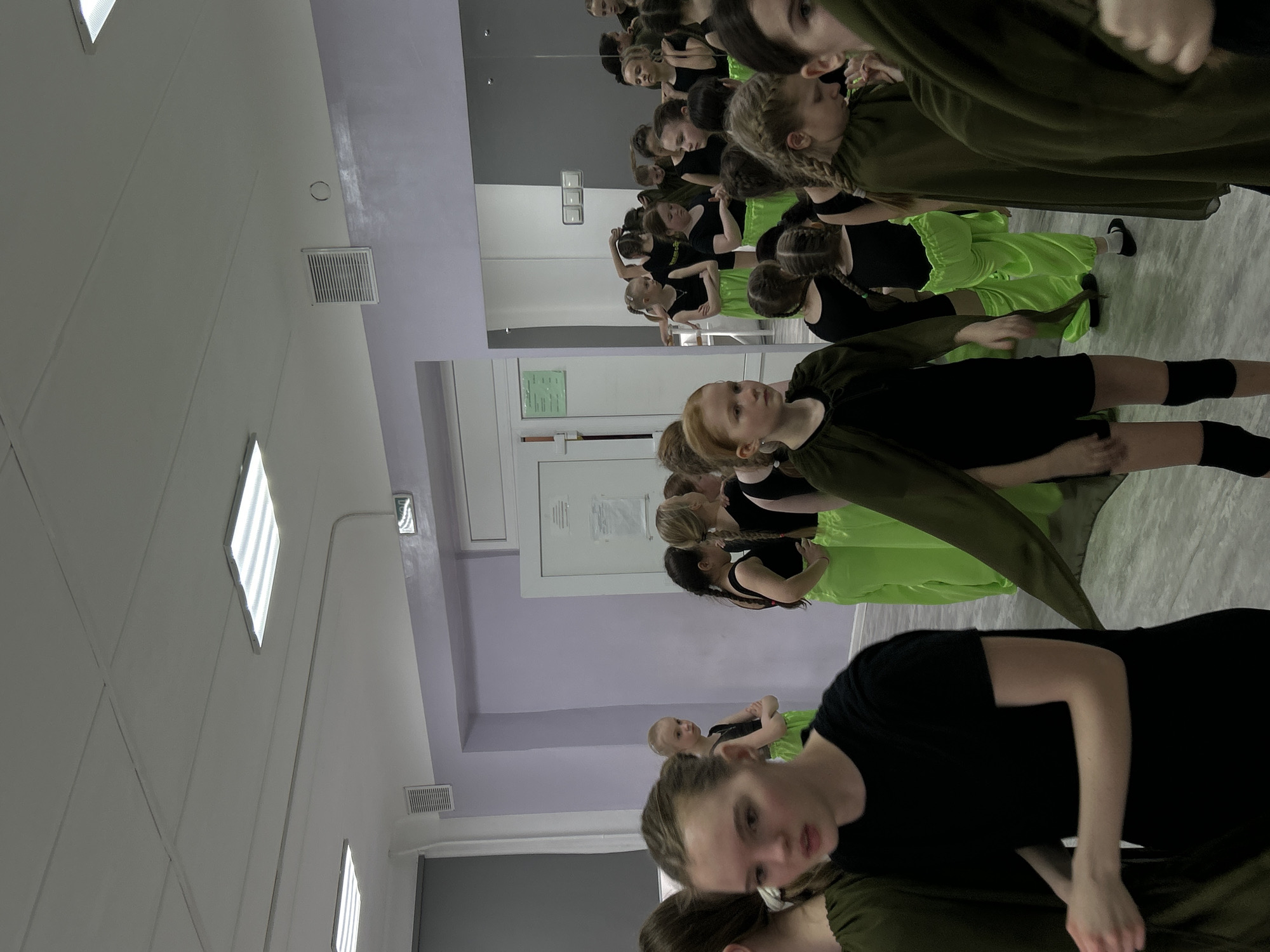 This media file captures a scene inside what appears to be a dance studio or rehearsal hall, likely in Chaikovsky, Russia. The setting features light-colored walls, primarily white, light grey, and a distinctive light lavender/purple section. Large mirrors line one wall, reflecting rows of individuals and extending the perceived depth of the room. The ceiling is equipped with several diamond-shaped recessed light fixtures and rectangular ventilation grates.
The primary subjects are numerous young girls and women, varying in age from possibly pre-teen to young adult, all dressed in practice or performance attire. They are arranged in several lines, suggesting a structured activity like a dance rehearsal or performance practice. Many wear black tops or full black outfits, often paired with bright, neon green flowing skirts or pants. Some individuals, particularly in the foreground and closer to the camera, are clad in dark olive green long-sleeved garments.
The activity taking place is a dance rehearsal or practice session, given the attire, the setting with mirrors, and the organized arrangement of the participants. Their expressions are generally focused or neutral. One girl in the immediate foreground, wearing a black top and olive green garment, looks towards the viewer with a thoughtful or attentive gaze.
Notable details include the artificial lighting from the ceiling fixtures, indicating an indoor setting. On a white door in the background, a paper notice with blurry Cyrillic text is visible. The overall atmosphere appears disciplined and purposeful, consistent with a dance training environment.