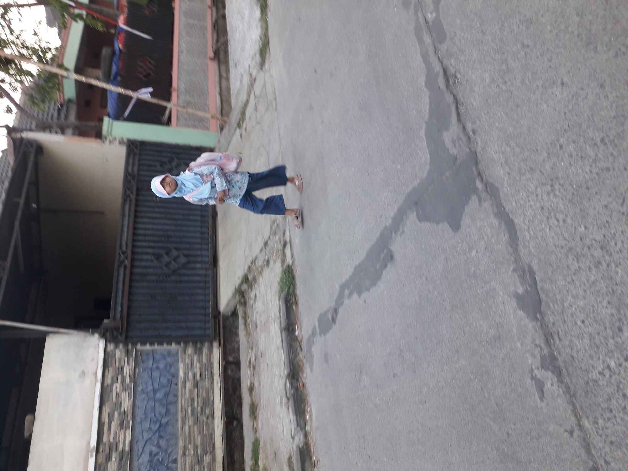 A young girl wearing a light blue hijab and a patterned long-sleeved shirt over blue pants walks on a paved road. She has a pink backpack on her back and is wearing sandals. The girl is walking away from the viewer and towards a house with a dark metal gate and a tiled facade. The house is located in Cankuang, Indonesia. The road is gray and has some cracks and water stains. There are some plants growing along the edge of the road. The lighting suggests it is late afternoon or early evening.