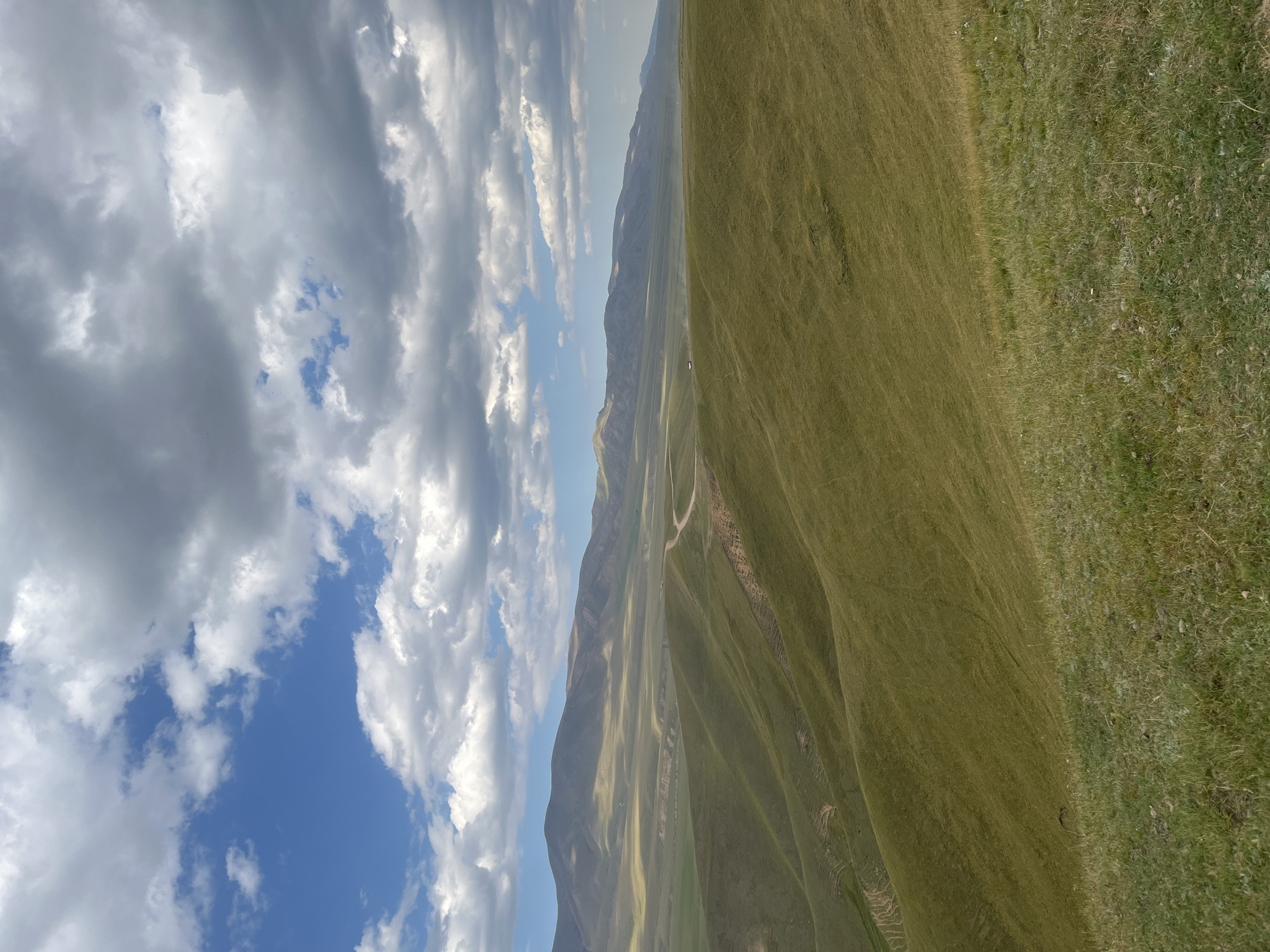 The image depicts a sweeping vista of rolling green hills under a vast, cloudy sky. The foreground is dominated by a grassy slope, with patches of sparser vegetation near the bottom. The midground reveals more undulating hills, some showing signs of terracing or erosion, with a dirt road winding its way through them. In the distance, a mountain range stretches across the horizon, bathed in a soft, hazy light. The sky is a mix of bright blue patches and dramatic, grey and white clouds. There are no discernible people or specific objects that would indicate a city location. The scene is characteristic of a rural, mountainous landscape.
