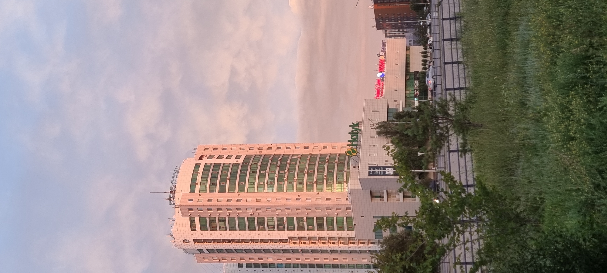 The image depicts a view of a modern cityscape in Aktobe, Kazakhstan, during what appears to be either sunrise or sunset, given the soft, pastel hues of the sky. Dominating the scene is a tall, slender, light pink apartment building with numerous windows, many of which are tinted green. The building has a curved facade and is topped with several antennas.
In front of this residential tower, and slightly to its right, is a lower, wider commercial building. This building features a prominent sign with green lettering that reads "Halyk," likely indicating a financial institution or bank. To the right of the "Halyk" sign, there are other illuminated signs in red and blue, suggesting a shopping center or entertainment complex. The facade of this commercial building is a lighter color, with geometric window patterns and balconies.
The foreground of the image is filled with lush, green vegetation, including trees and tall grass, indicating a park or undeveloped area near the roadside. A multi-lane road is visible behind the vegetation, with a dark metal fence separating it from the grassy area. Several cars are visible on the road, moving in the same direction. To the far right of the commercial building, another building is visible, this one appearing to be a taller, darker structure. The overall impression is one of urban development juxtaposed with natural elements.