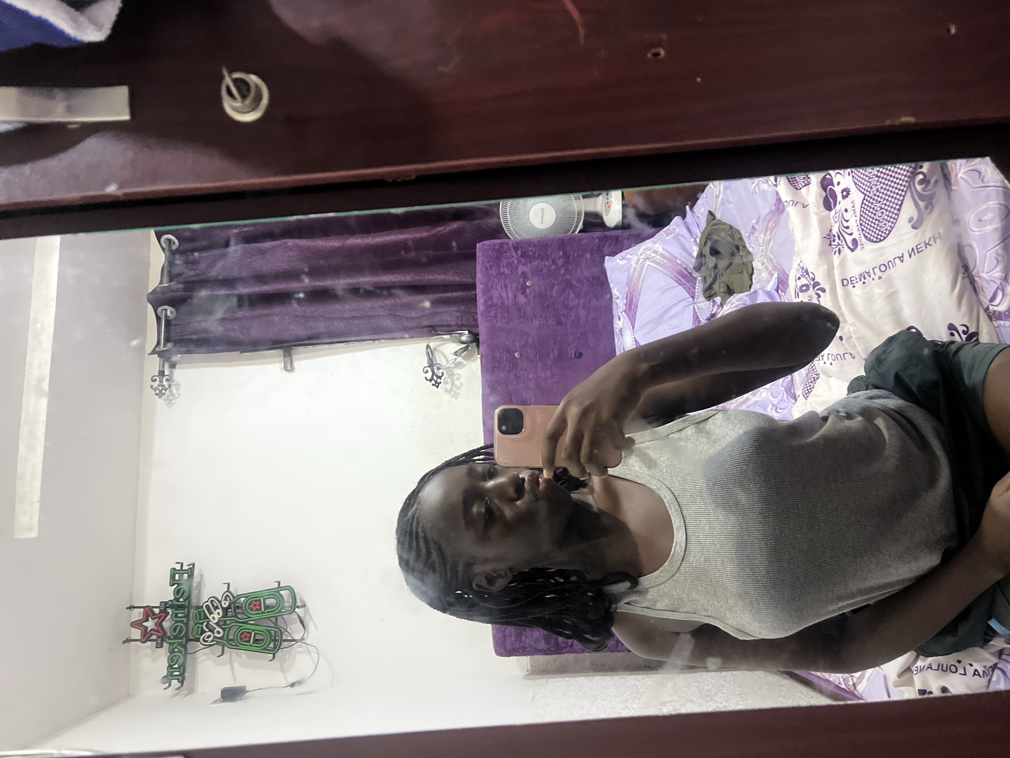A woman takes a mirror selfie in what appears to be a bedroom. The room features a bed with patterned sheets, a purple headboard, and curtains. A small electric fan is visible in the background. A neon sign, possibly advertising Heineken, is mounted on the wall. The location is likely a private residence in Abuja, Nigeria, though this cannot be confirmed with certainty. The subject is taking a picture of herself in a mirror.