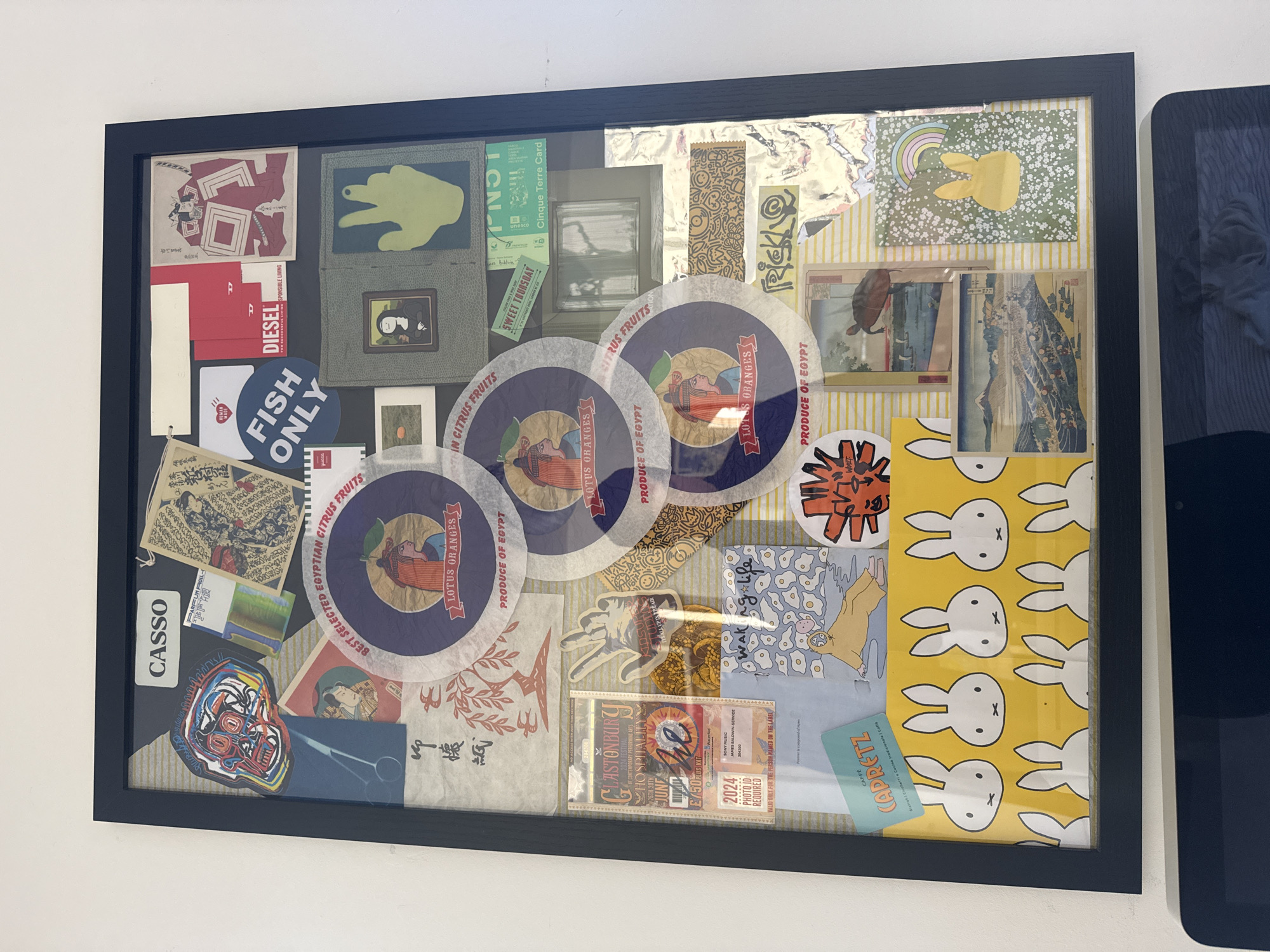 The image shows a framed collage of various items. Key elements include stickers with the words "DIESEL", "FISH ONLY", and "CASSO"; circular labels with "LOTUS ORANGES" and "PRODUCE OF EGYPT"; a ticket to "GLASTONBURY HOSPITALITY" for £750; and a card for "CAFFE CAPRETZ". Other items include patches, illustrations, and patterned paper. The frame is black and rectangular. The collage is mounted on a white wall, with a portion of a black electronic tablet or device visible to the right of the frame. The location cannot be determined from the image.